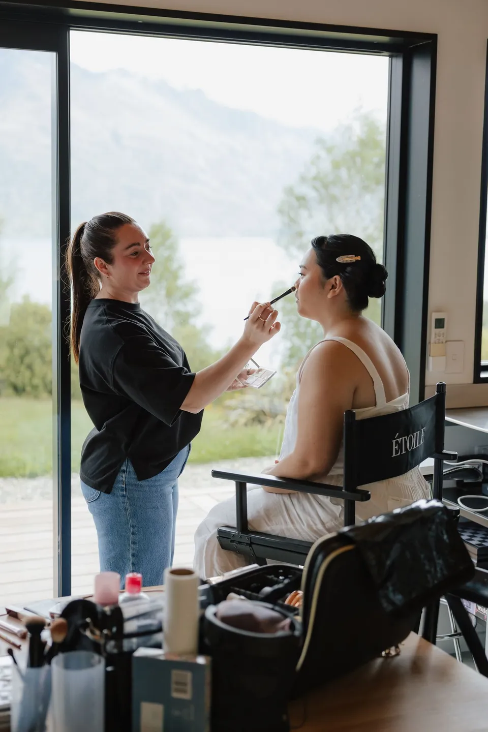 Jaeda having her bridal makeup applied beside a window overlooking the lake and mountains, at their alpine Airbnb in Ben Lomond, Queenstown, New Zealand, for an Elopement Collective Queenstown elopement. Photograph by Bulb Creative.