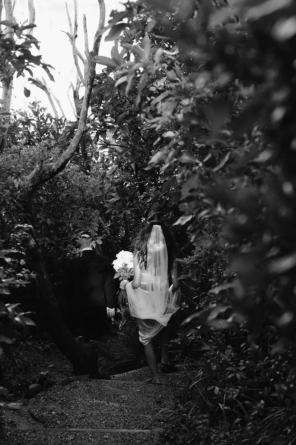Black-and-white photograph of Rachel carrying her bouquet down a leafy coastal path, with flowers by Fifth Avenue Florist. At Whites Beach, Broken Head, near Byron Bay, for an Elopement Collective elopement. Photograph by Jason Lucas.