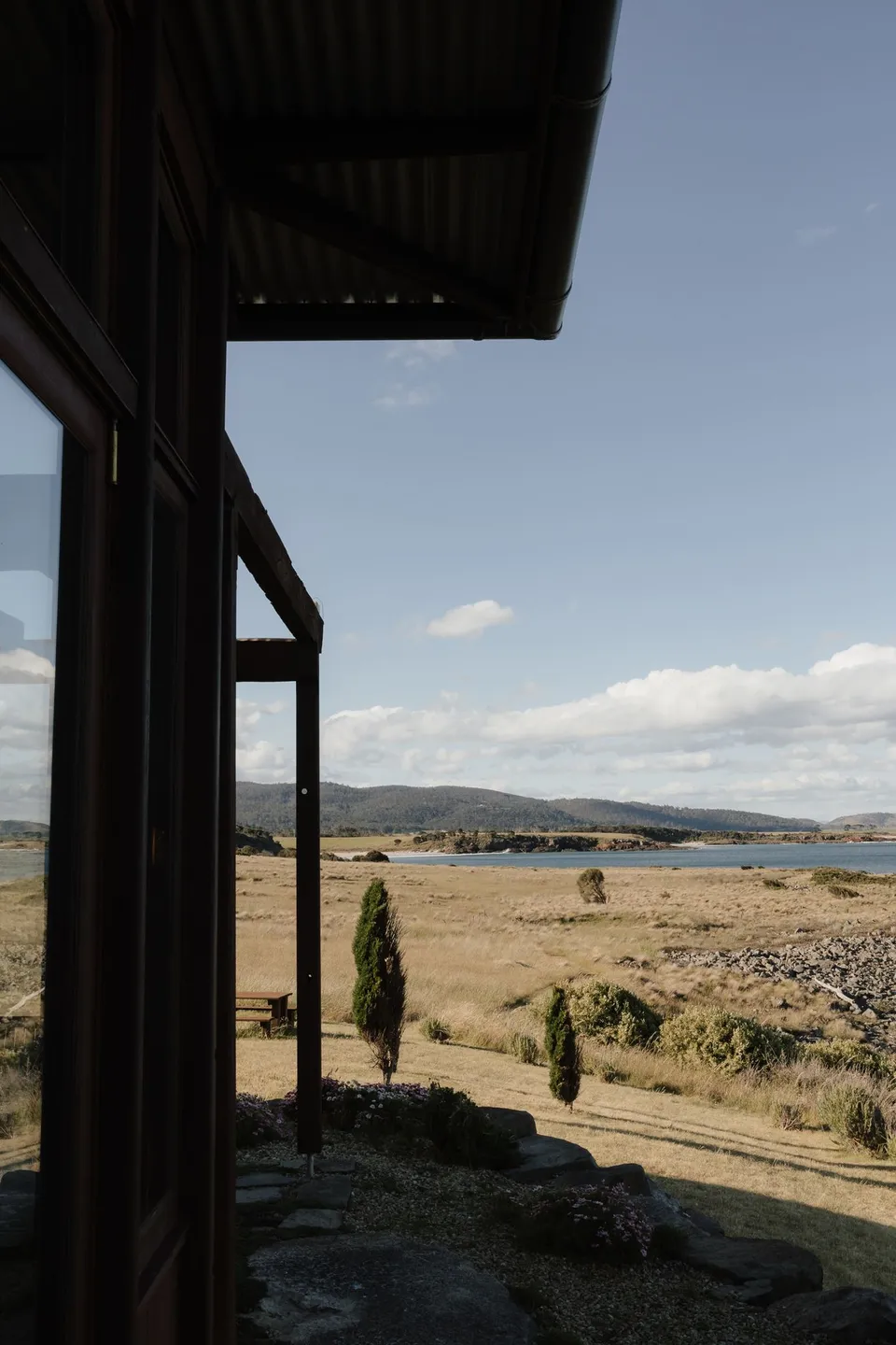 An open timber-framed window in the stone house, looking across golden grassland towards the coast, at Thalia Haven on the East Coast of Tasmania, for an Elopement Collective elopement. Photograph by Bulb Creative.