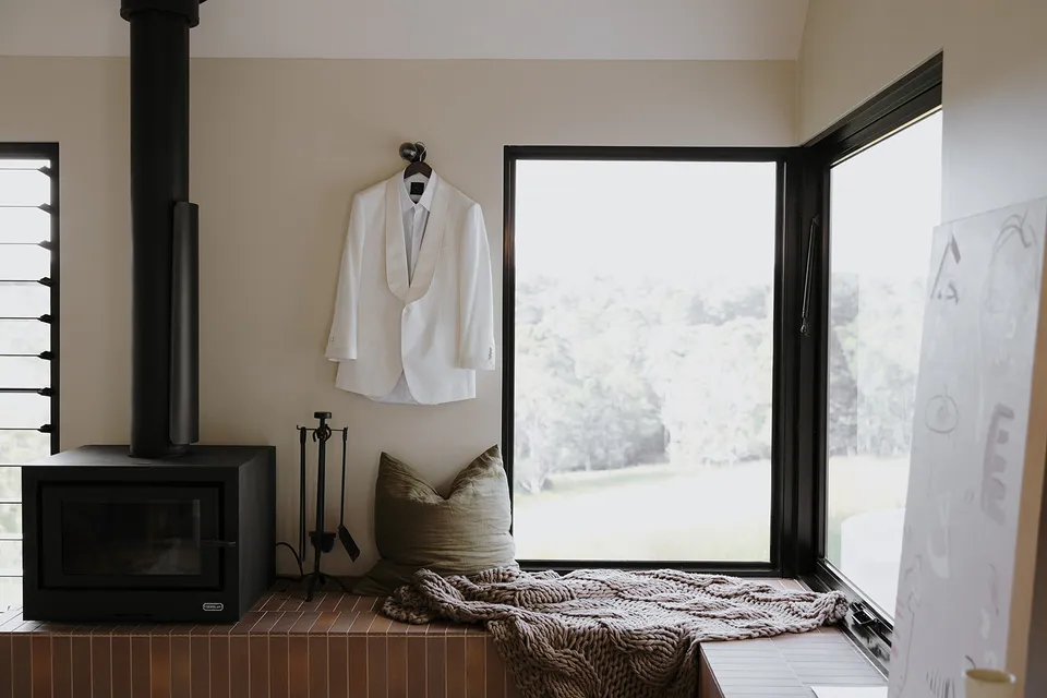 Greg’s white Norfords dinner jacket hanging beside the cabin fireplace and an open window seat, at Roundhill Retreat in Obi Obi, Sunshine Coast Hinterland, Queensland, for an Elopement Collective elopement. Photo by George Bowden.