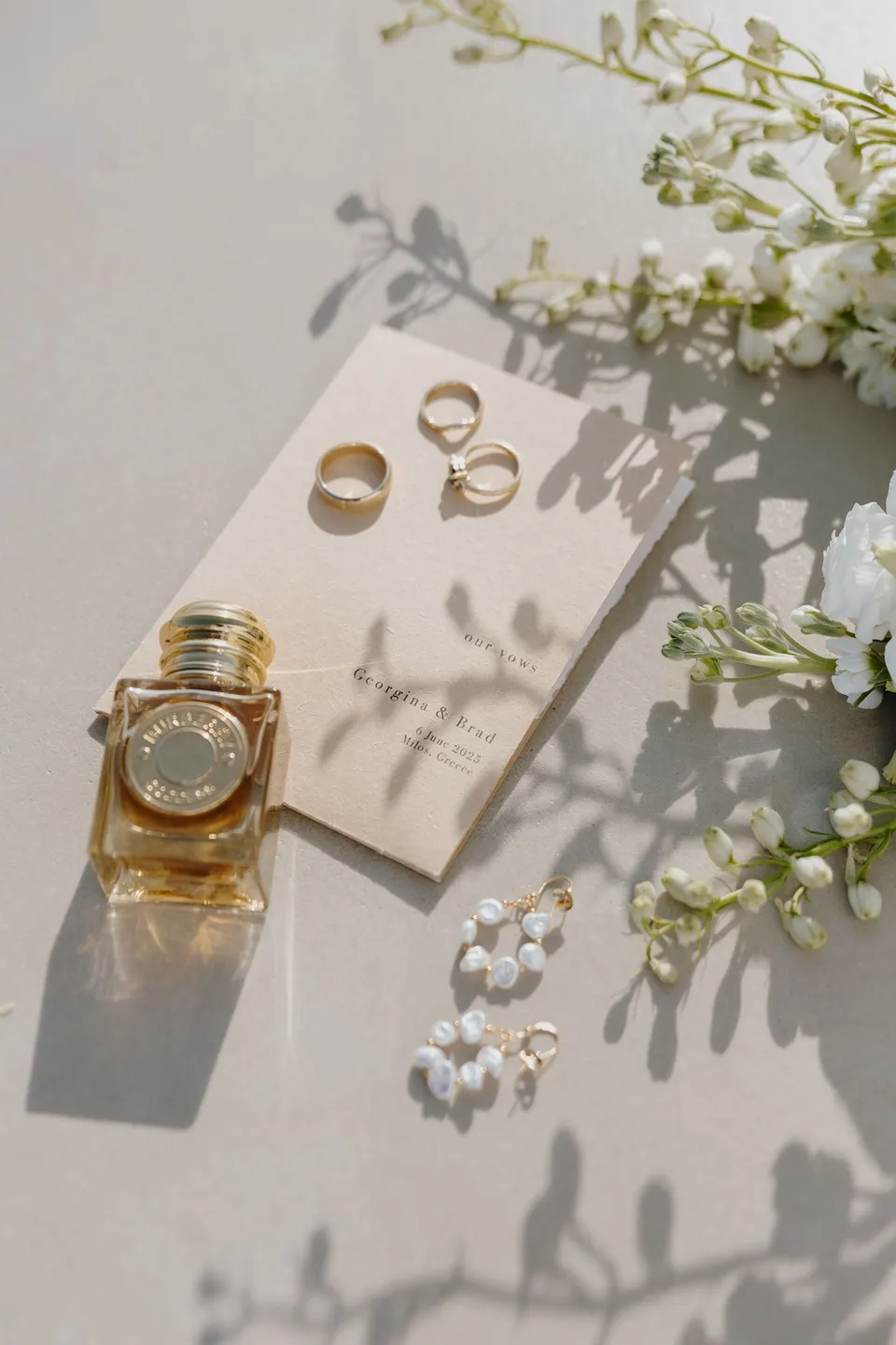Wedding rings on Georgina and Brad’s personalised vow book, beside perfume, pearl hoop earrings and white flowers by Giasemi, at Adriana Luxury Villas on Milos, Greece, during an Elopement Collective elopement. Photograph by George Bowden.