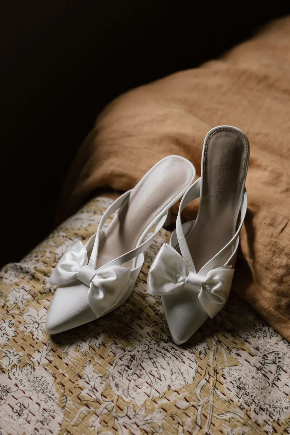 White pointed wedding shoes with bow details resting on a patterned bedspread. At Cudgera Creek in northern New South Wales, for an Elopement Collective elopement. Photograph by Bulb Creative.