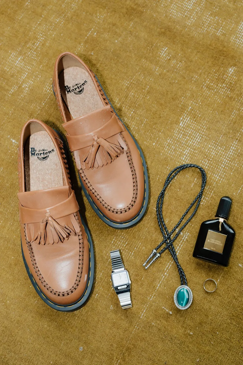 Adrian’s tan tassel loafers, turquoise bolo tie, watch and cologne arranged on a gold velvet chair, at their alpine Airbnb in Ben Lomond, Queenstown, New Zealand, for an Elopement Collective Queenstown elopement. Photograph by Bulb Creative.