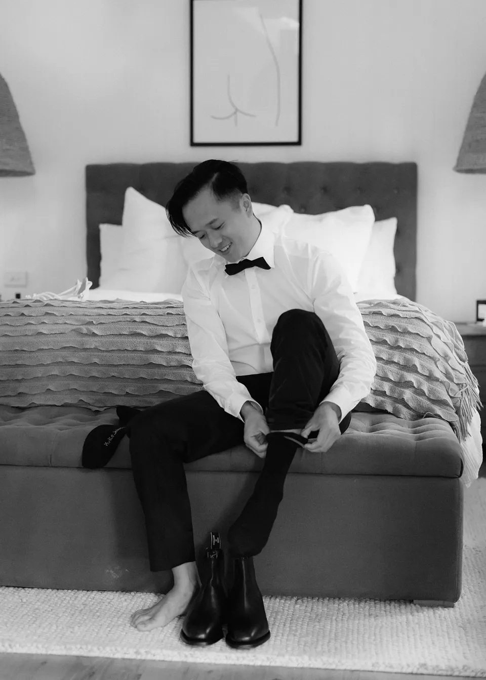 Black-and-white photograph of Jonathan getting ready beside the bed. At Fig Tree Villa in Ewingsdale, Byron Bay, for an Elopement Collective elopement. Photograph by Bulb Creative.