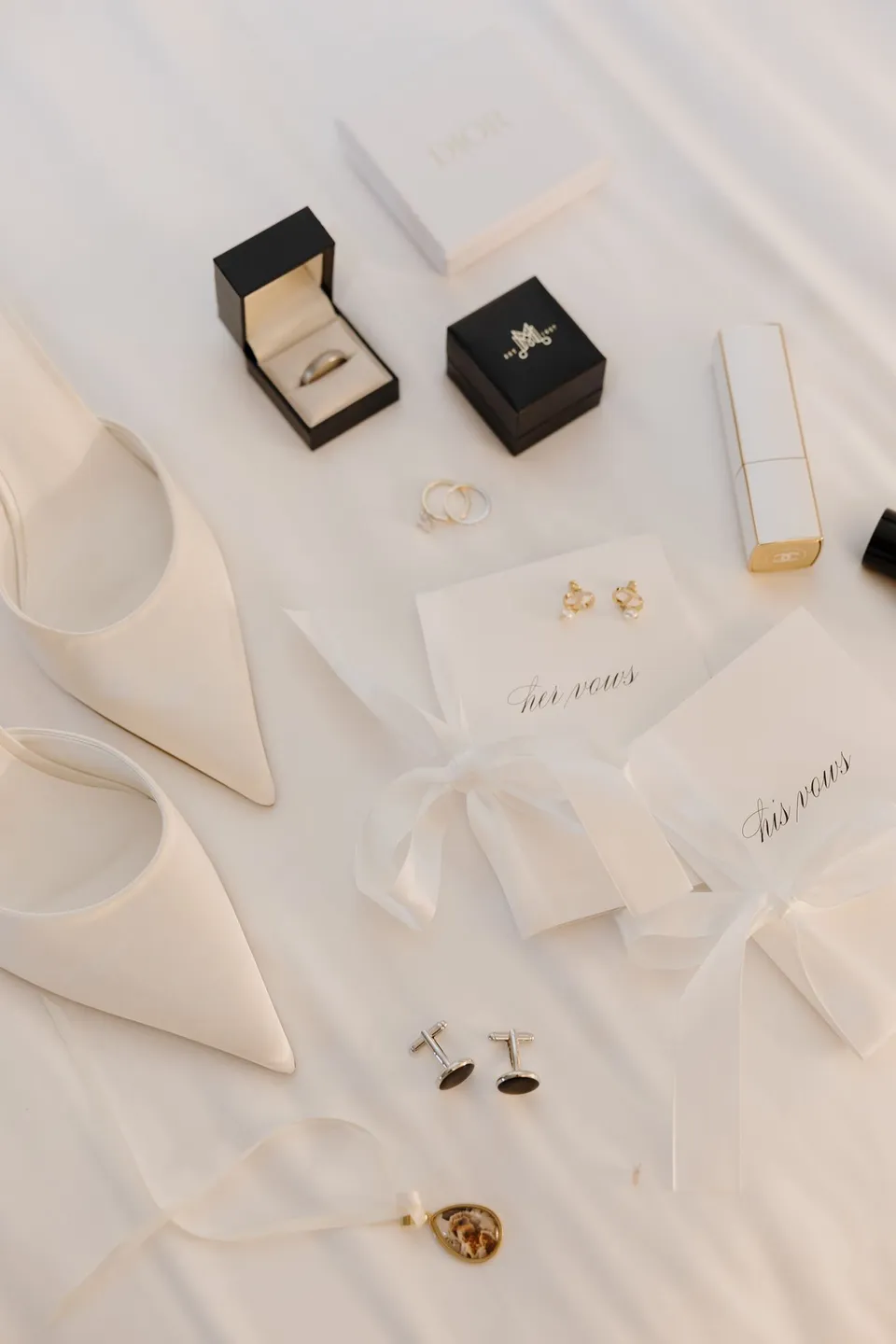 Wedding rings, ribbon-tied vow books, pointed white shoes, earrings, cufflinks and perfume arranged on a white bedspread, at Passalacqua in Moltrasio, Lake Como, Italy, for an Elopement Collective elopement. Photo by George Bowden.