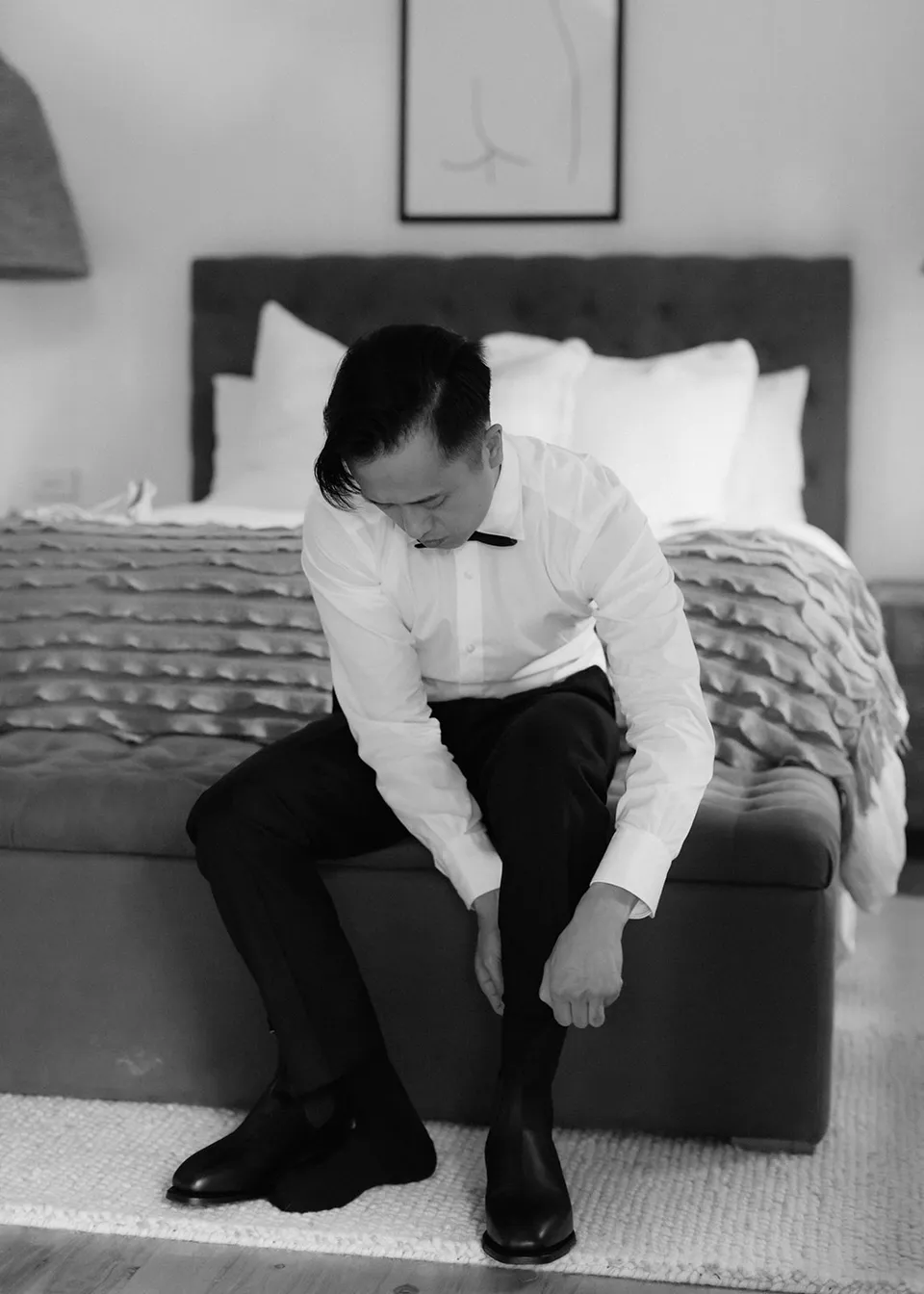 Black-and-white photograph of Jonathan leaning forward to fasten his boots. At Fig Tree Villa in Ewingsdale, Byron Bay, for an Elopement Collective elopement. Photograph by Bulb Creative.