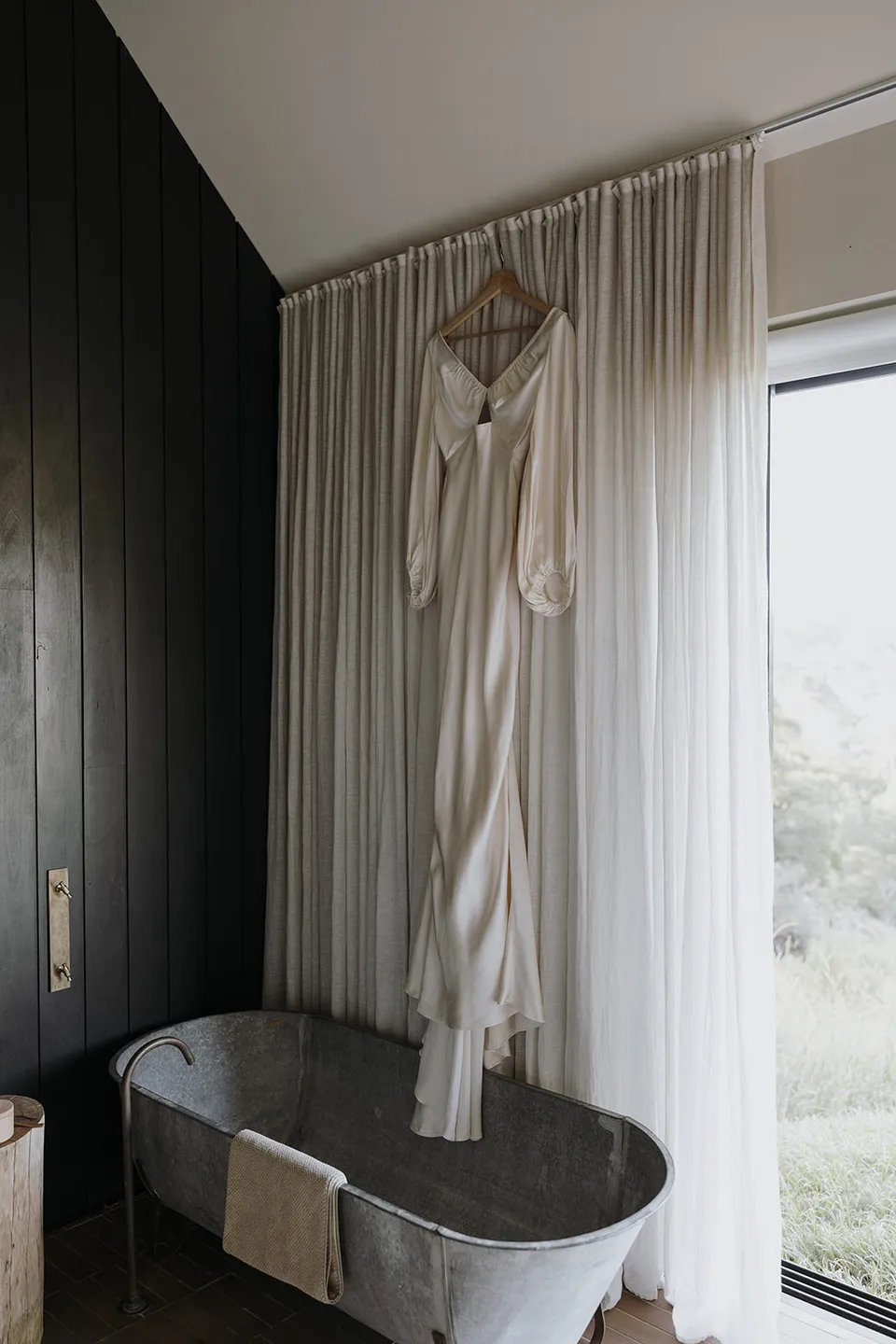 Gabrielle’s ivory Shona Joy dress hanging above a freestanding bath beside the cabin window, at Roundhill Retreat in Obi Obi, Sunshine Coast Hinterland, Queensland, for an Elopement Collective elopement. Photo by George Bowden.