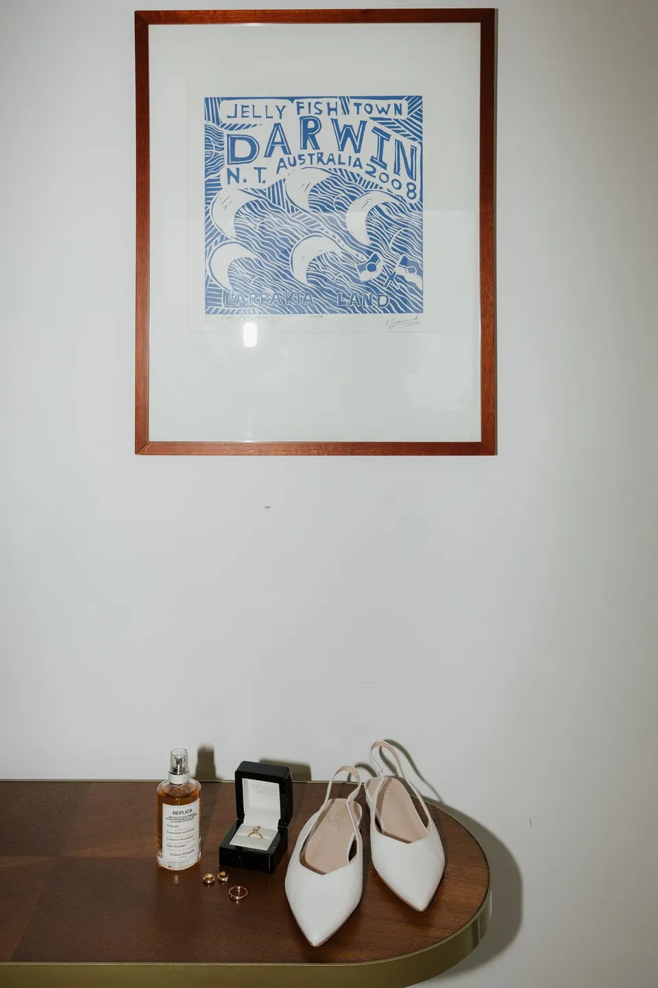 Jaeda’s white pointed bridal shoes and perfume displayed on a timber table beneath a framed print, at their alpine Airbnb in Ben Lomond, Queenstown, New Zealand, for an Elopement Collective Queenstown elopement. Photograph by Bulb Creative.