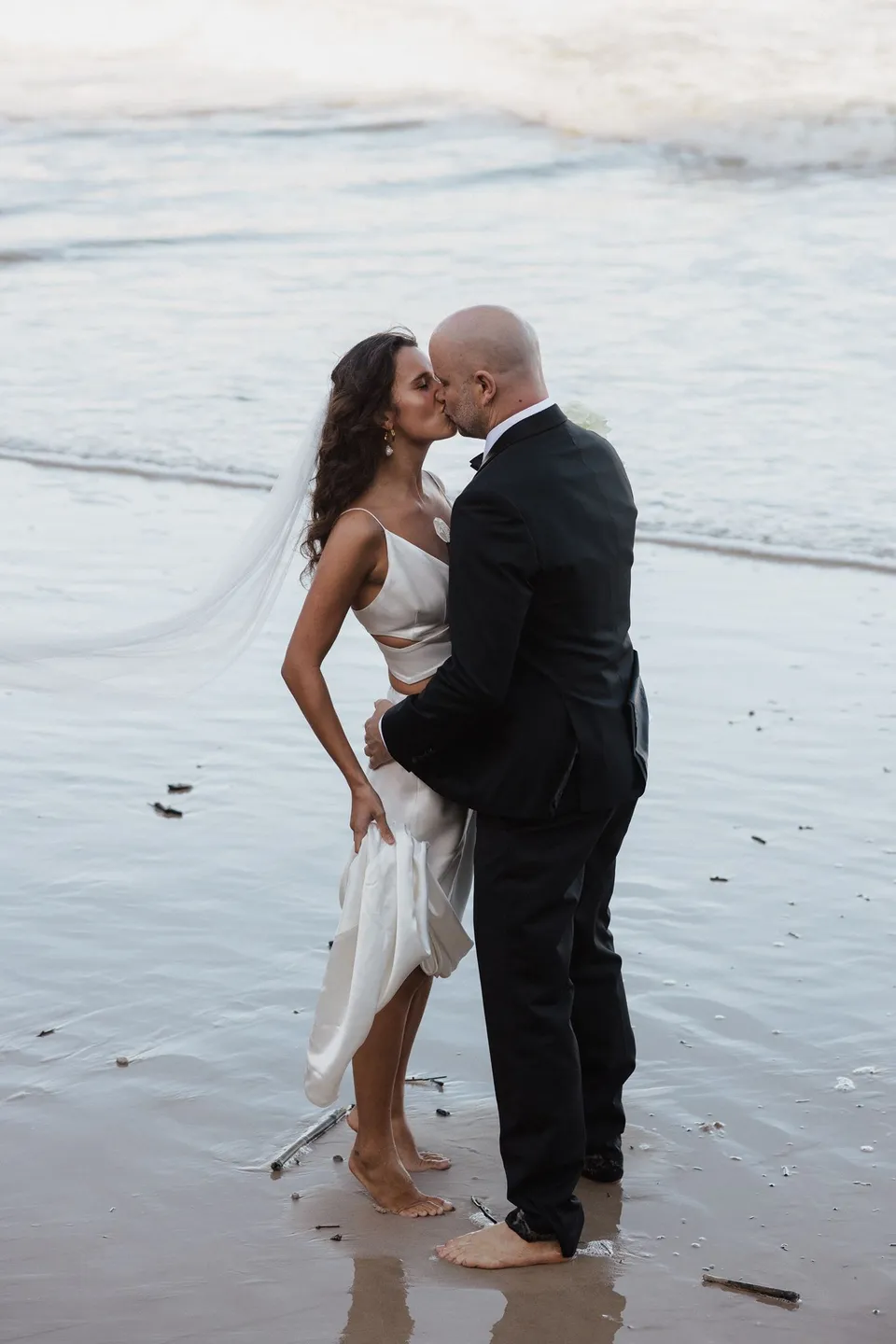 Rachel and Dane kissing barefoot on the wet sand beside the ocean. At Whites Beach, Broken Head, near Byron Bay, for an Elopement Collective elopement. Photograph by Jason Lucas.