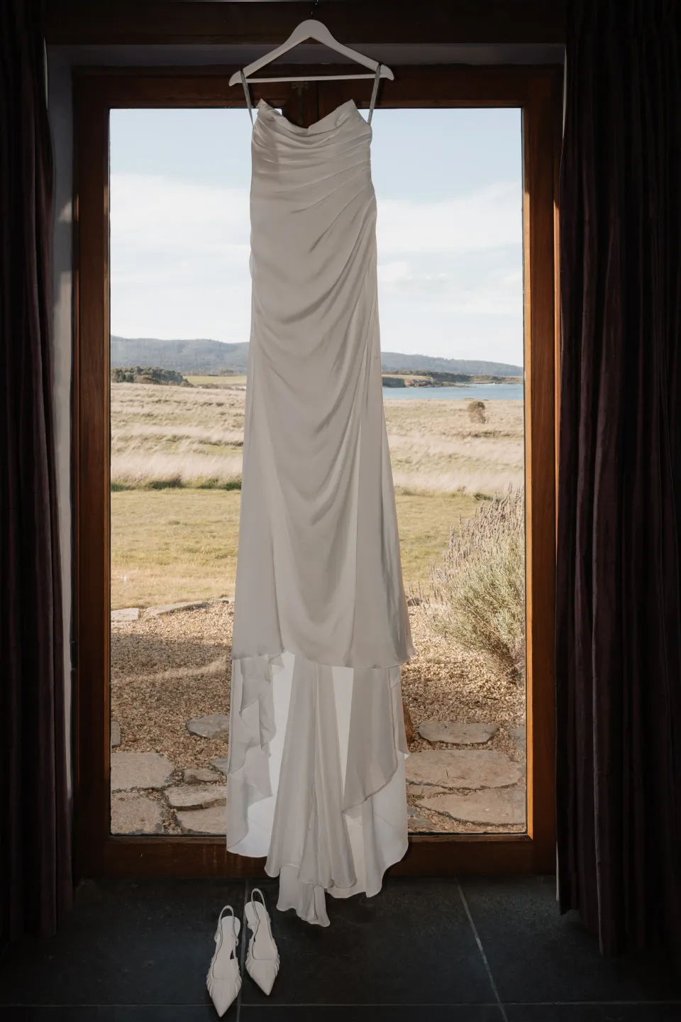 Dani’s draped white wedding dress hanging above her shoes in a dark timber doorway, with the coast outside, at Thalia Haven on Tasmania’s east coast, for an Elopement Collective elopement. Photograph by Bulb Creative.