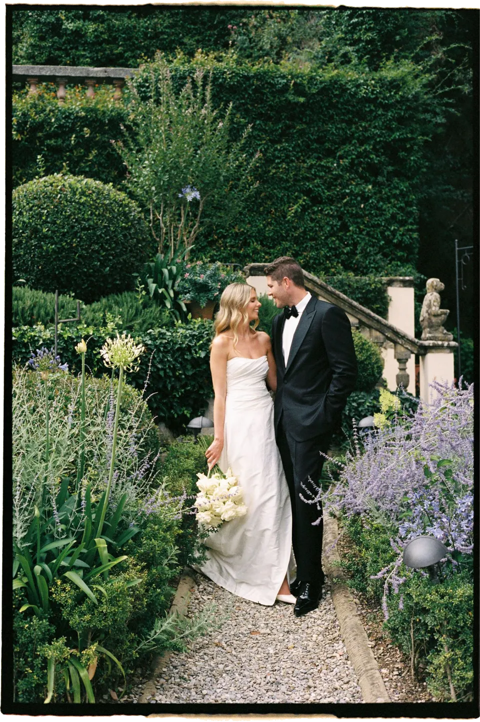 Holly and Matthew kissing on a garden path surrounded by purple flowers, with her white bouquet by Le Francesche Flower Designers, at Villa San Michele in Fiesole above Florence, Tuscany, Italy, for an Elopement Collective elopement. Film photograph by Khara from House of Lucie.