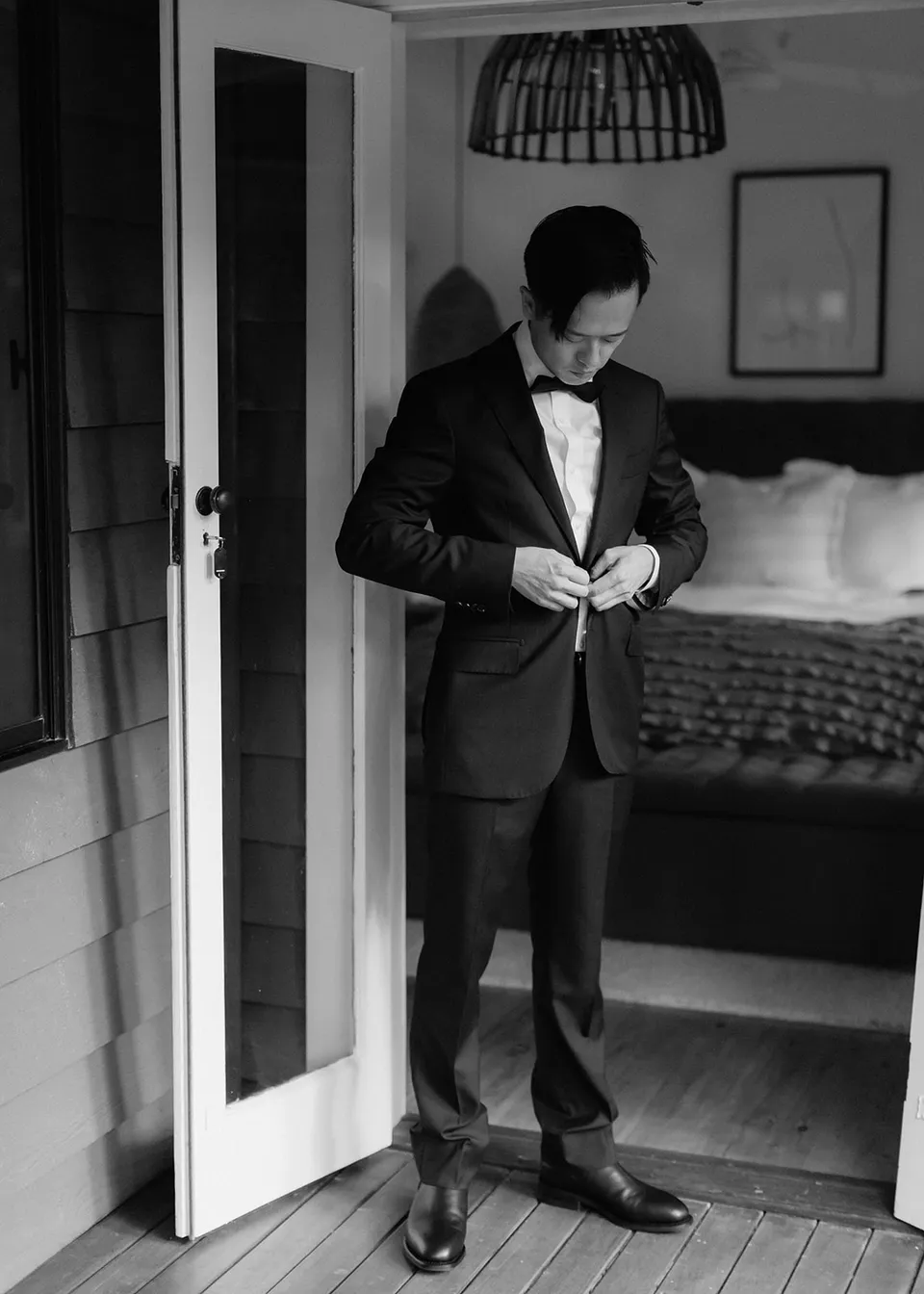 Black-and-white full-length portrait of Jonathan fastening his jacket in the doorway. At Fig Tree Villa in Ewingsdale, Byron Bay, for an Elopement Collective elopement. Photograph by Bulb Creative.