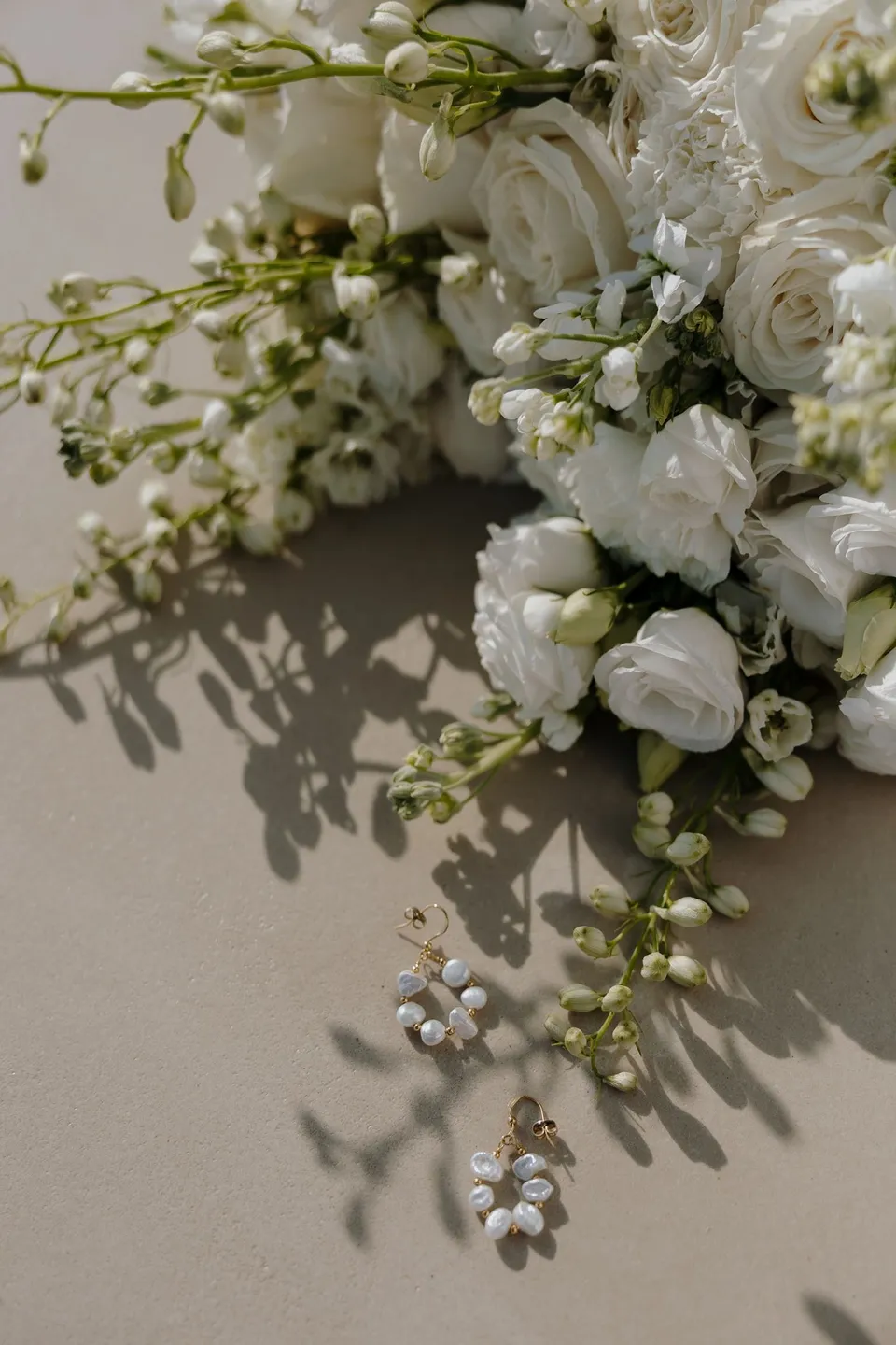 A white rose bouquet by Giasemi with green stems, beside pearl hoop earrings on a pale surface, at Adriana Luxury Villas on Milos, Greece, during an Elopement Collective elopement. Photograph by George Bowden.