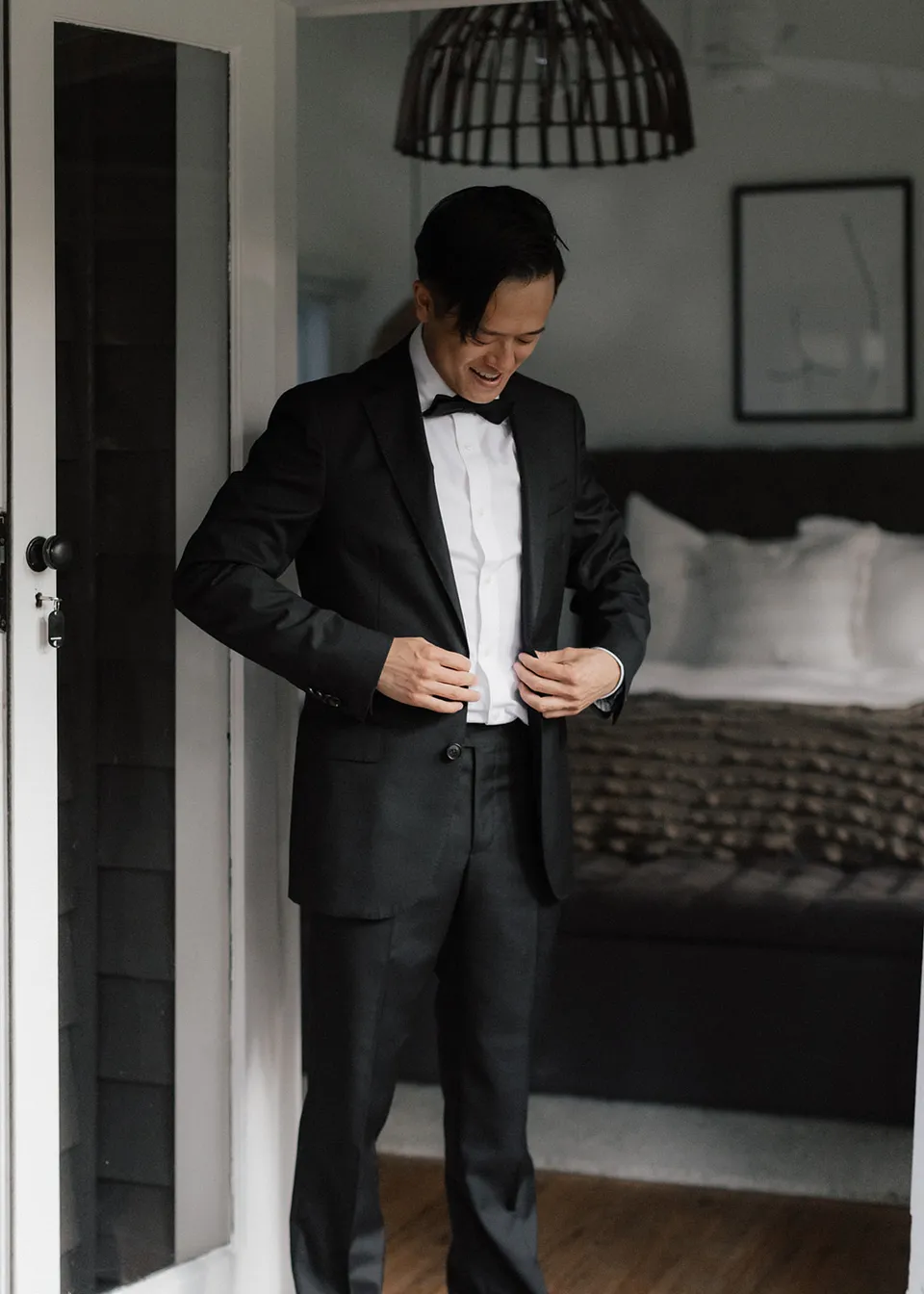 Jonathan smiling while adjusting his suit jacket beside the bedroom door. At Fig Tree Villa in Ewingsdale, Byron Bay, for an Elopement Collective elopement. Photograph by Bulb Creative.