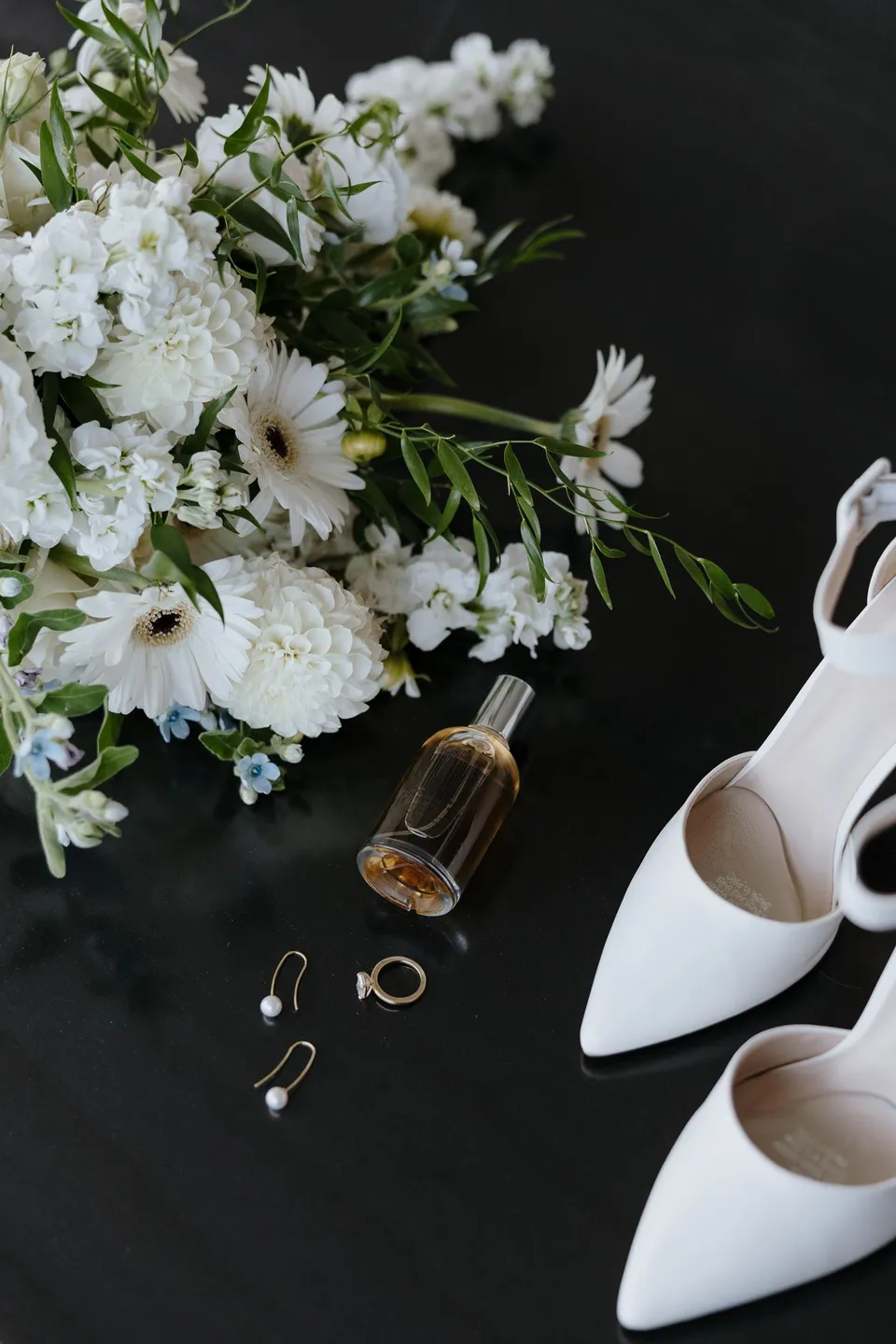 White bridal shoes, rings, earrings and perfume beside a white bouquet by Orange Blossom, at their Airbnb in Fernhill, Queenstown, New Zealand, for an Elopement Collective Queenstown elopement. Photo by George Bowden.