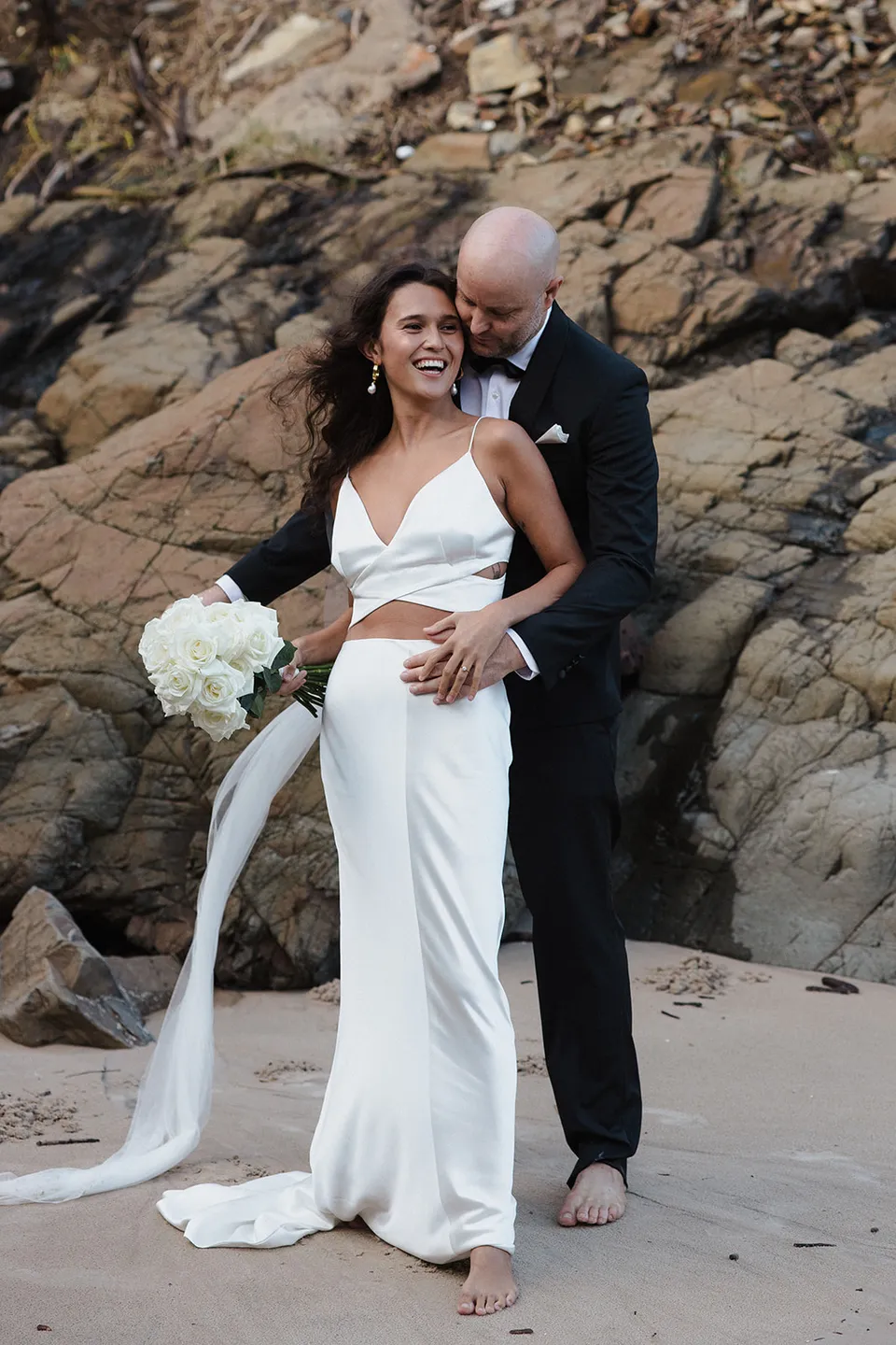 Dane embracing Rachel from behind beside the rock face as she holds her white bouquet, with flowers by Fifth Avenue Florist. At Whites Beach, Broken Head, near Byron Bay, for an Elopement Collective elopement. Photograph by Jason Lucas.