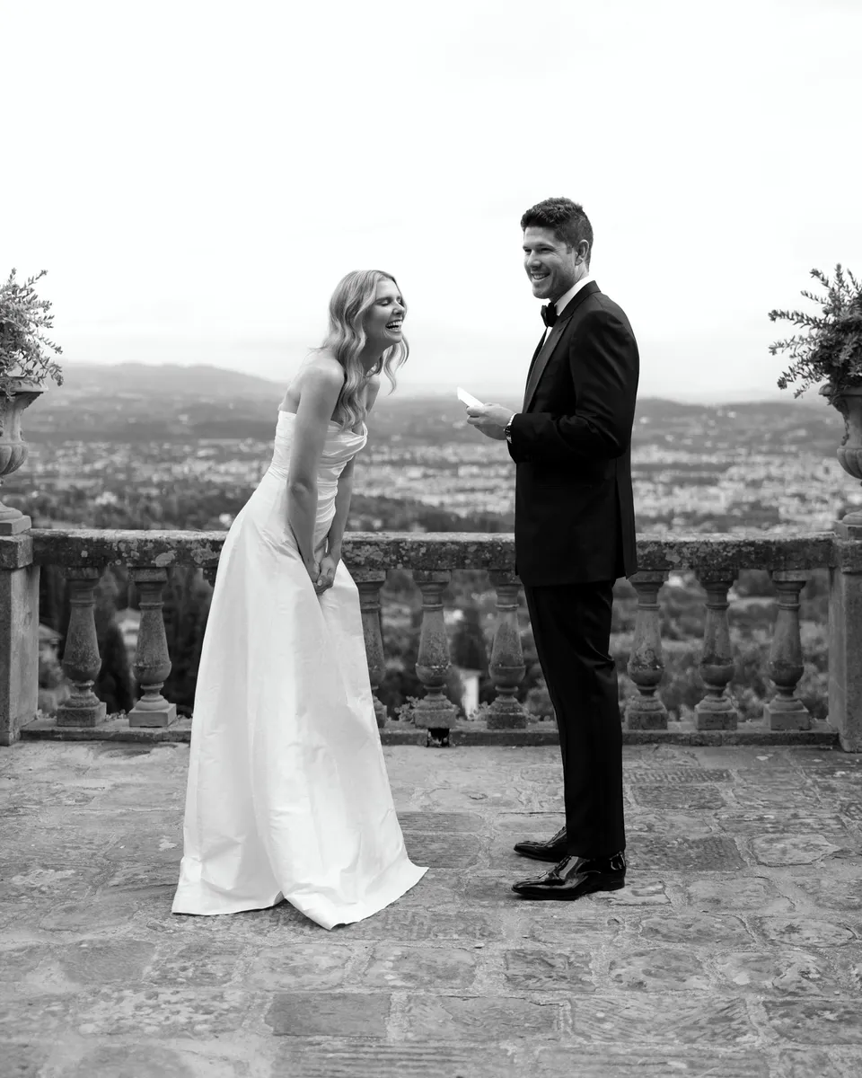 Black-and-white photograph of Matthew smiling with his vow card as Holly listens, at Villa San Michele in Fiesole above Florence, Tuscany, Italy, for an Elopement Collective elopement. Digital photograph by Khara from House of Lucie.