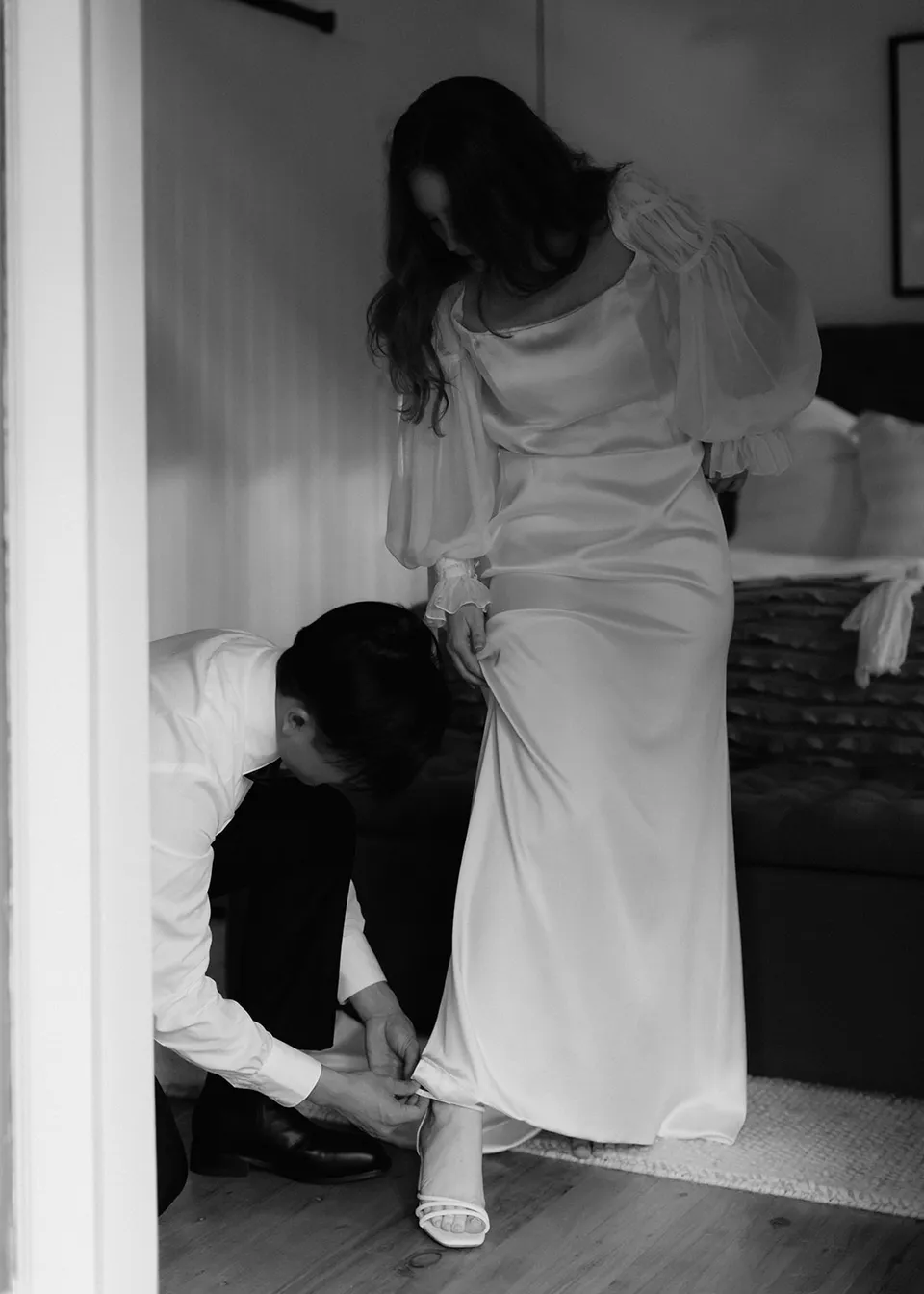 Black-and-white photograph of Jonathan kneeling to help Elyse with her shoes. At Fig Tree Villa in Ewingsdale, Byron Bay, for an Elopement Collective elopement. Photograph by Bulb Creative.