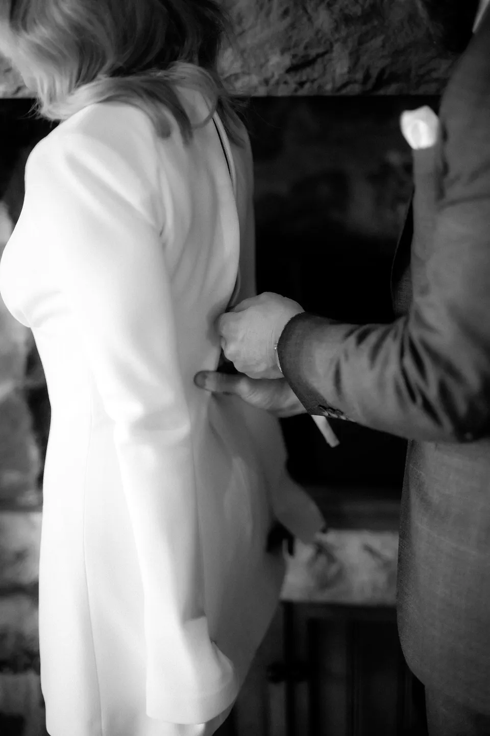 Black and white close-up of Russell fastening the back of Kaitlyn’s long-sleeved short wedding dress. At Piermont Retreat on Tasmania’s east coast, for an Elopement Collective elopement. Photograph by Omar of By The Wilde.
