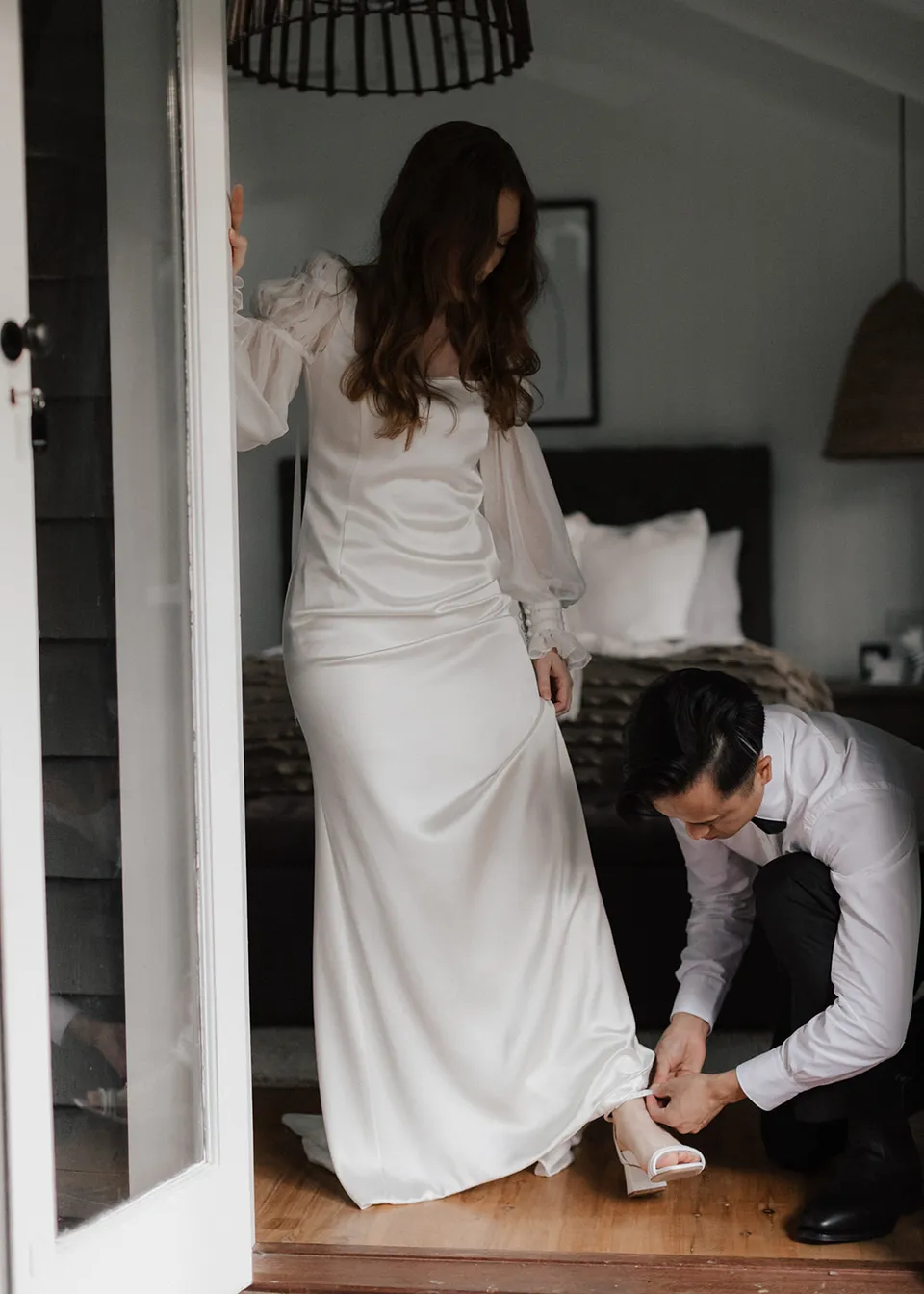 Elyse standing in her dress while Jonathan helps with her shoes. At Fig Tree Villa in Ewingsdale, Byron Bay, for an Elopement Collective elopement. Photograph by Bulb Creative.