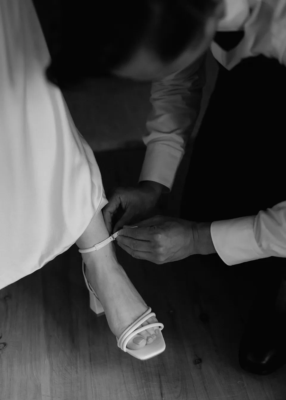 Black-and-white close-up of Jonathan fastening Elyse's shoe strap. At Fig Tree Villa in Ewingsdale, Byron Bay, for an Elopement Collective elopement. Photograph by Bulb Creative.