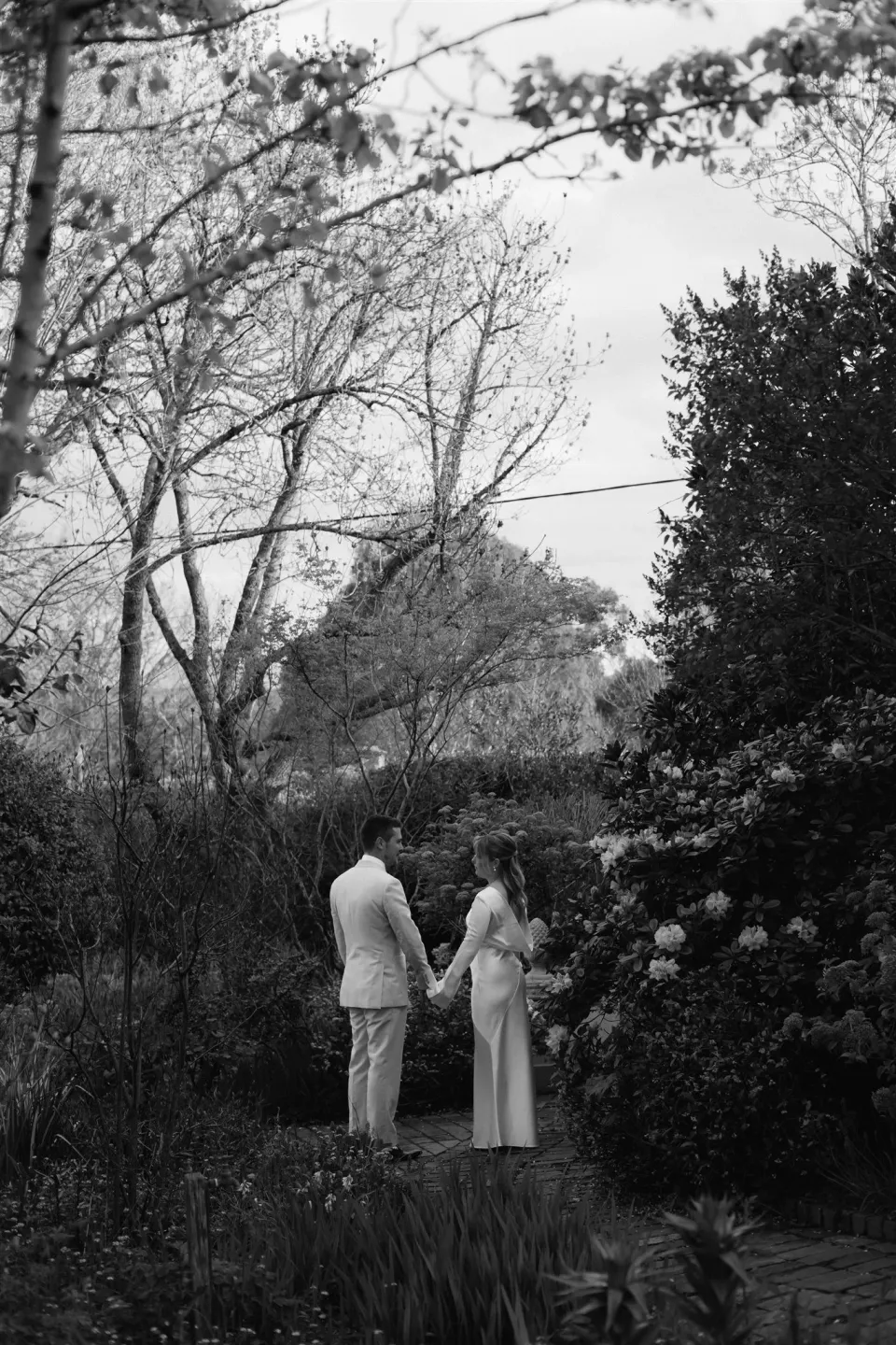 Black-and-white photograph of Maddy and Cameron holding hands beneath bare branches in the garden, at The Mayor’s Residence in Daylesford, Victoria, for an Elopement Collective elopement. Photo by Bulb Creative.