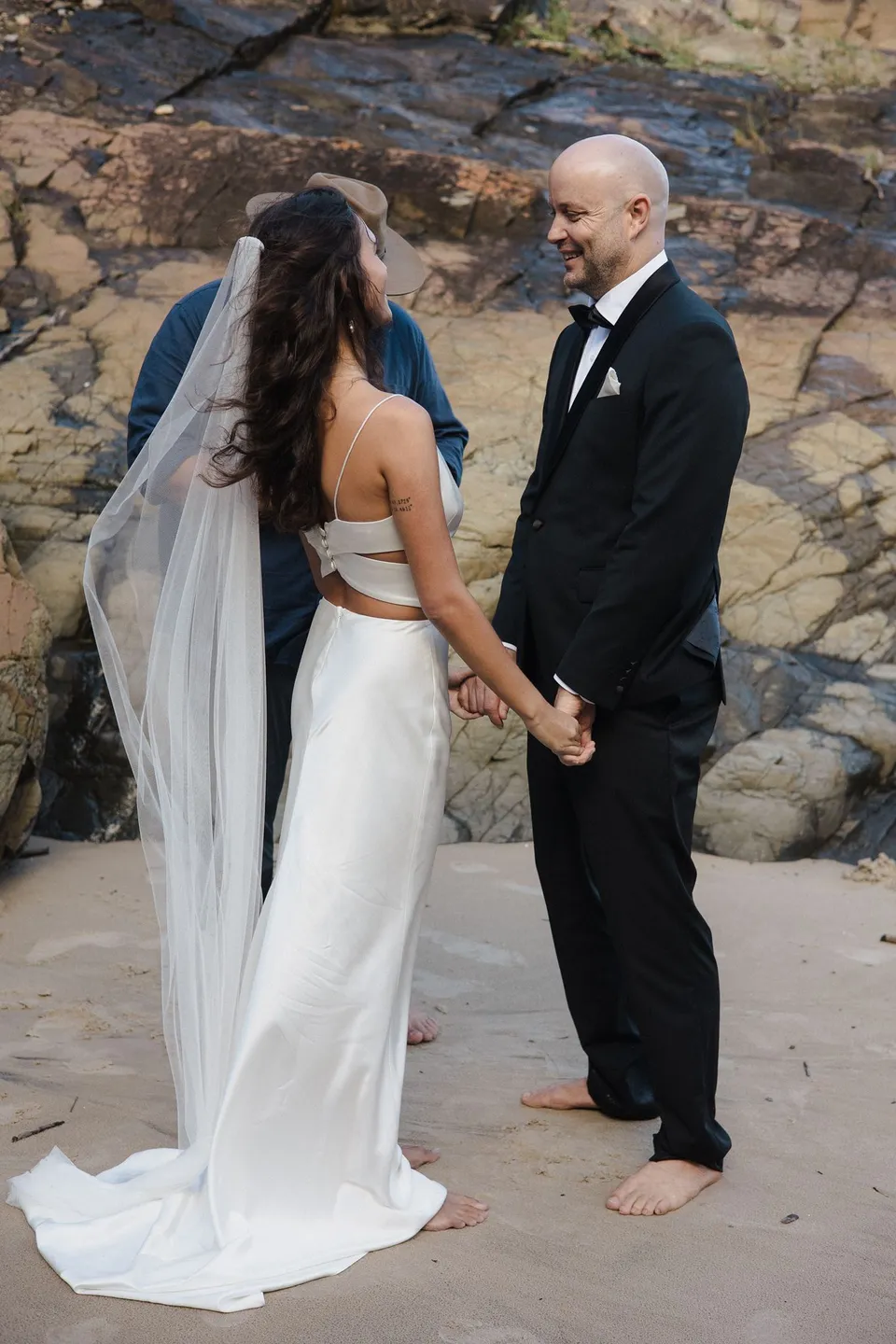 Rachel and Dane holding hands barefoot on the sand during their ceremony. At Whites Beach, Broken Head, near Byron Bay, for an Elopement Collective elopement. Photograph by Jason Lucas.