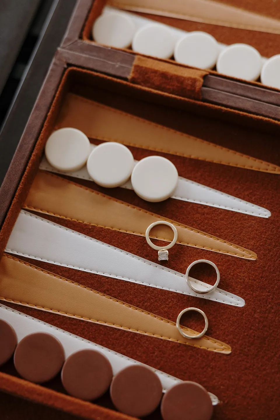 Wedding and engagement rings resting inside an open wooden backgammon board, at their Airbnb in Fernhill, Queenstown, New Zealand, for an Elopement Collective Queenstown elopement. Photo by George Bowden.