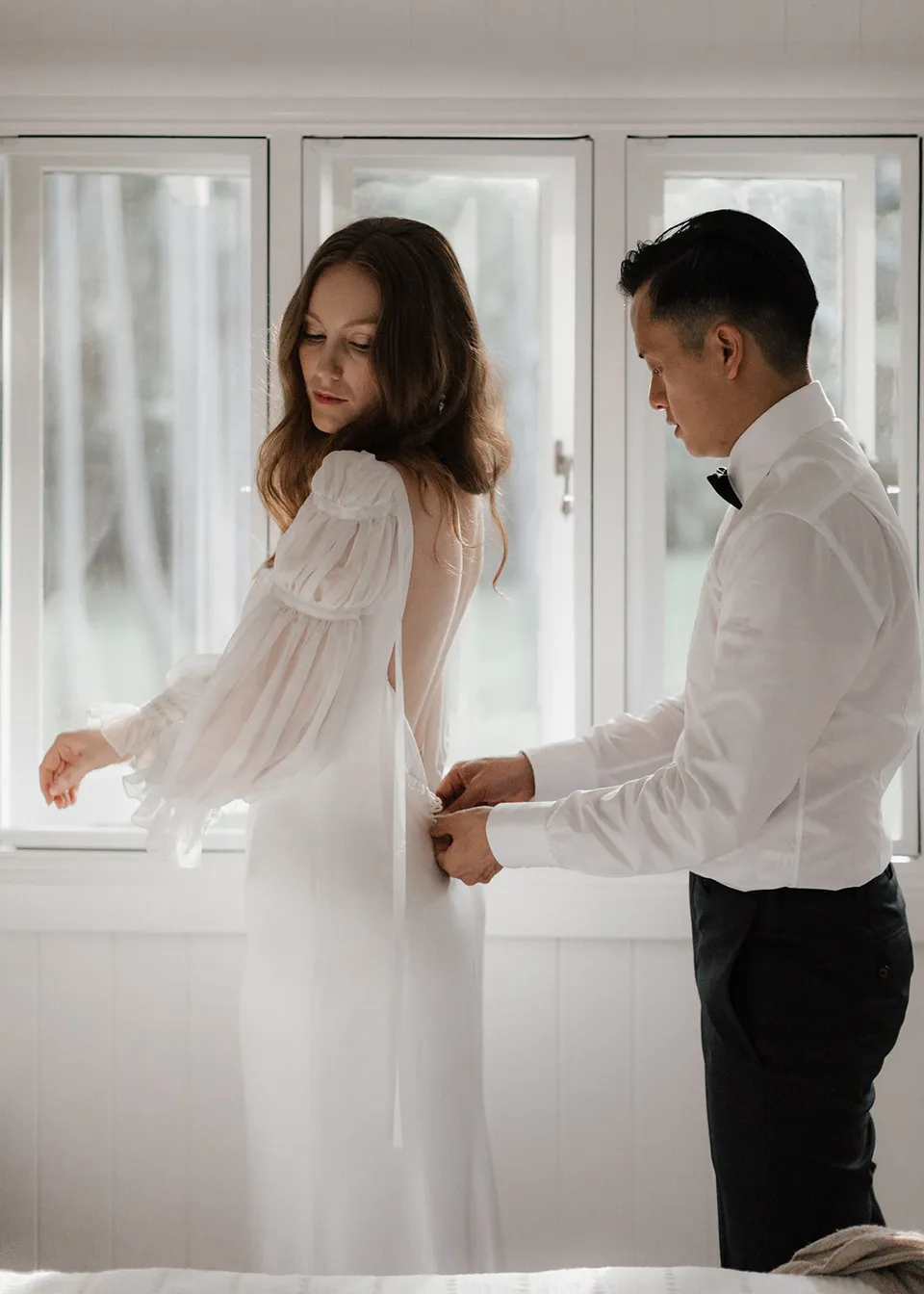 Elyse looking back at Jonathan as he fastens her dress. At Fig Tree Villa in Ewingsdale, Byron Bay, for an Elopement Collective elopement. Photograph by Bulb Creative.