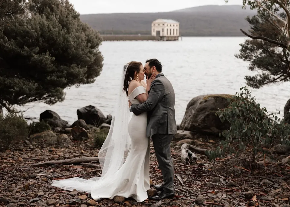 Sarah and Jono kissing on the rocky lakeshore with the Pumphouse building behind them, at Pumphouse Point, Lake St Clair, Tasmania, for an Elopement Collective elopement. Photo by Bulb Creative.