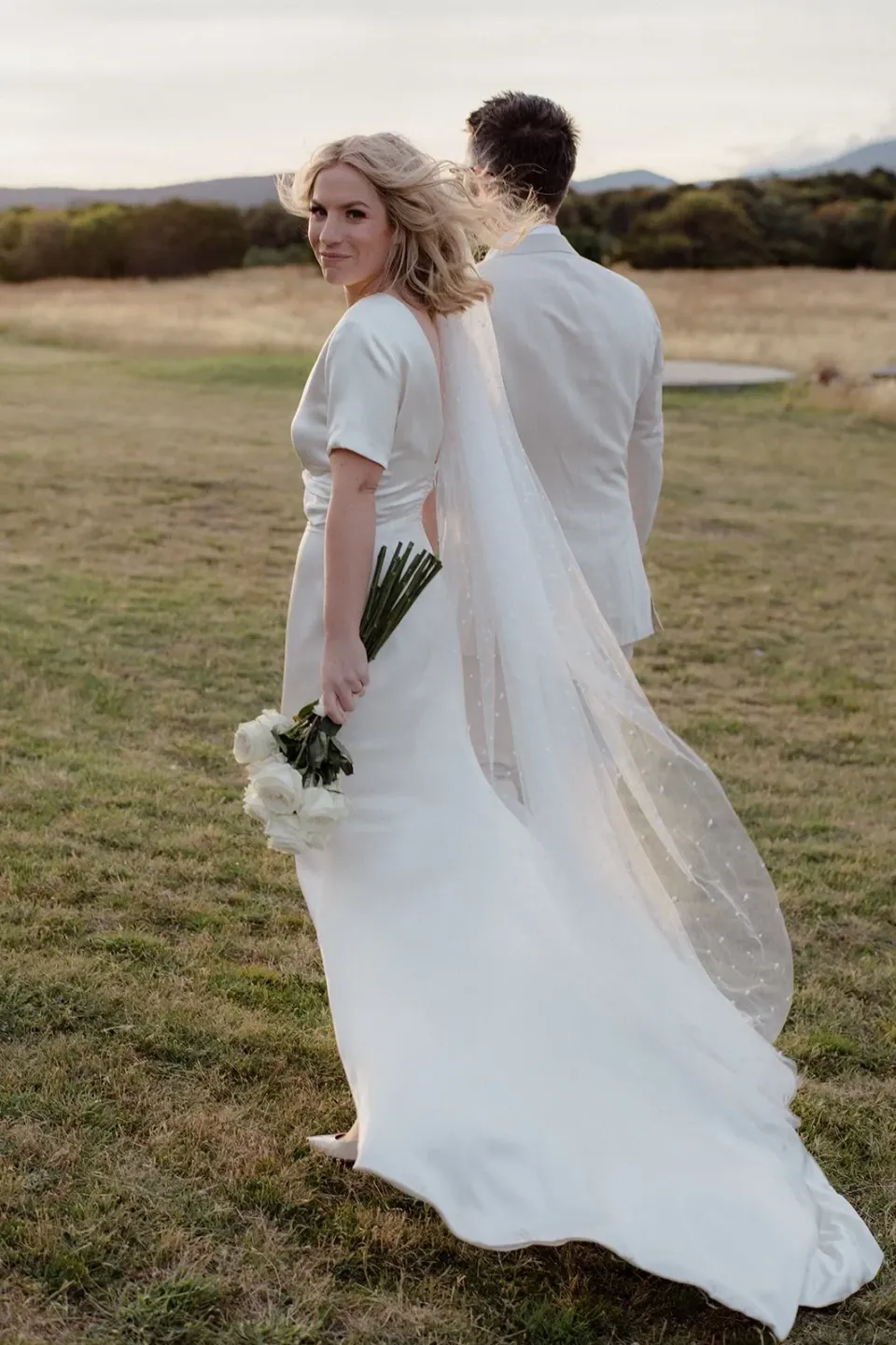 A bride turns towards the camera as her veil trails across an open lawn.