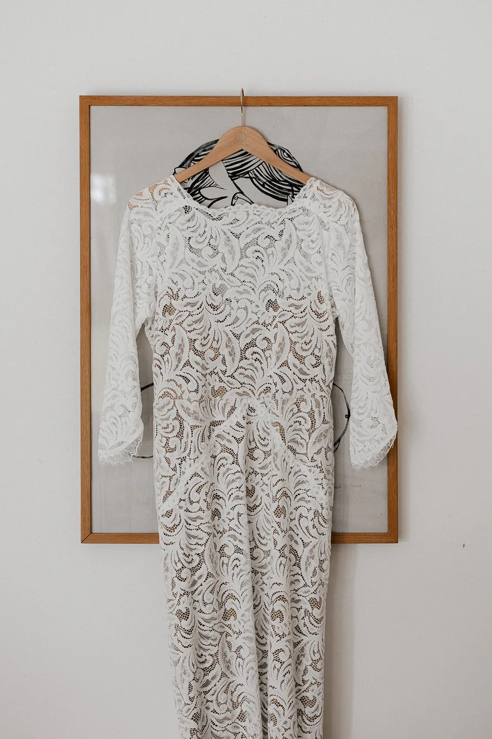Laura’s long-sleeved lace wedding dress hanging against a framed artwork, at Grounds of Cabarita in Cabarita Beach, NSW, for an Elopement Collective elopement. Photo by Bulb Creative.