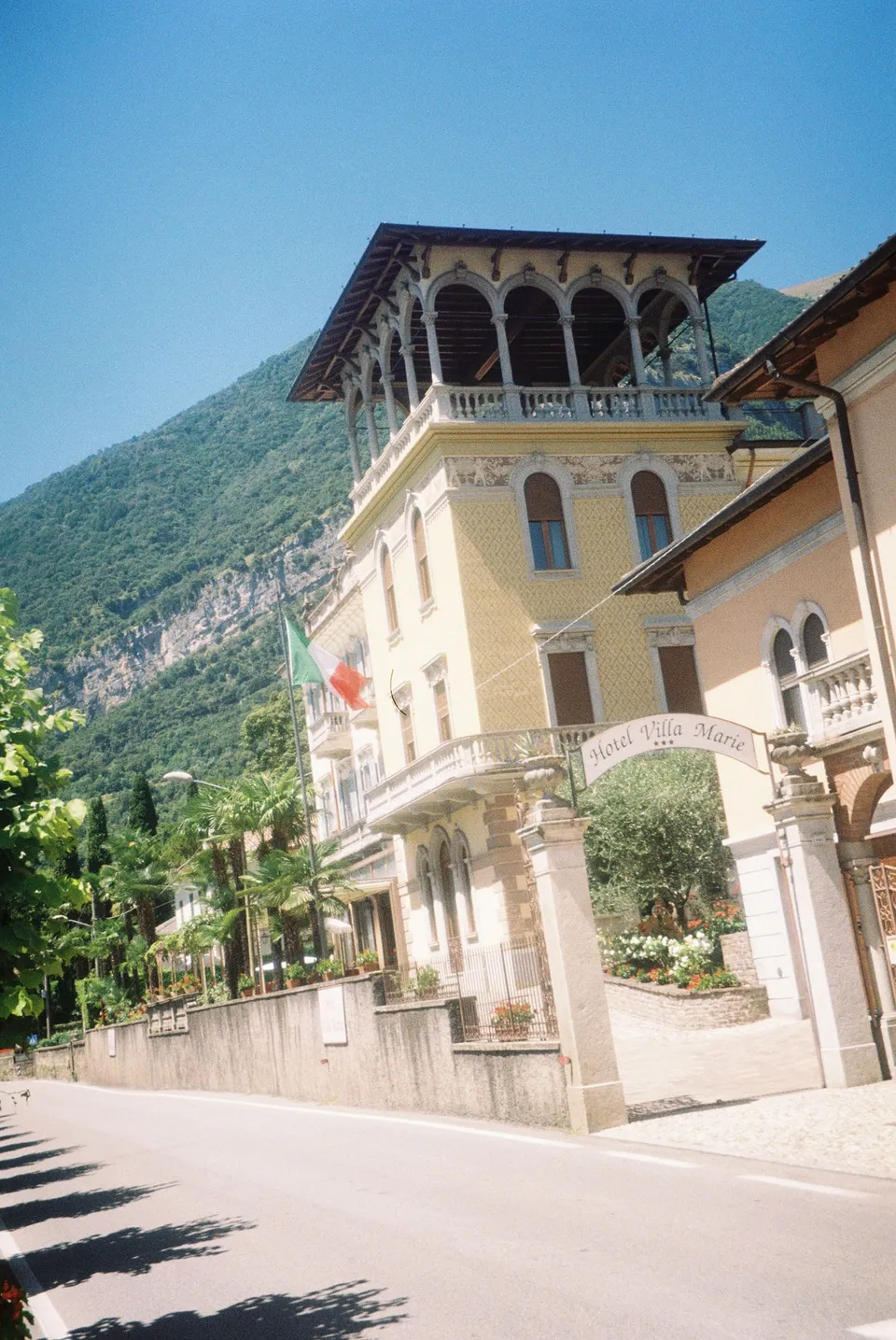 Hotel Villa Marie’s pale facade, balconies and palm trees beside the road in Tremezzo, at Lake Como in Lombardy, Italy, for an Elopement Collective elopement. Photograph by George Bowden.