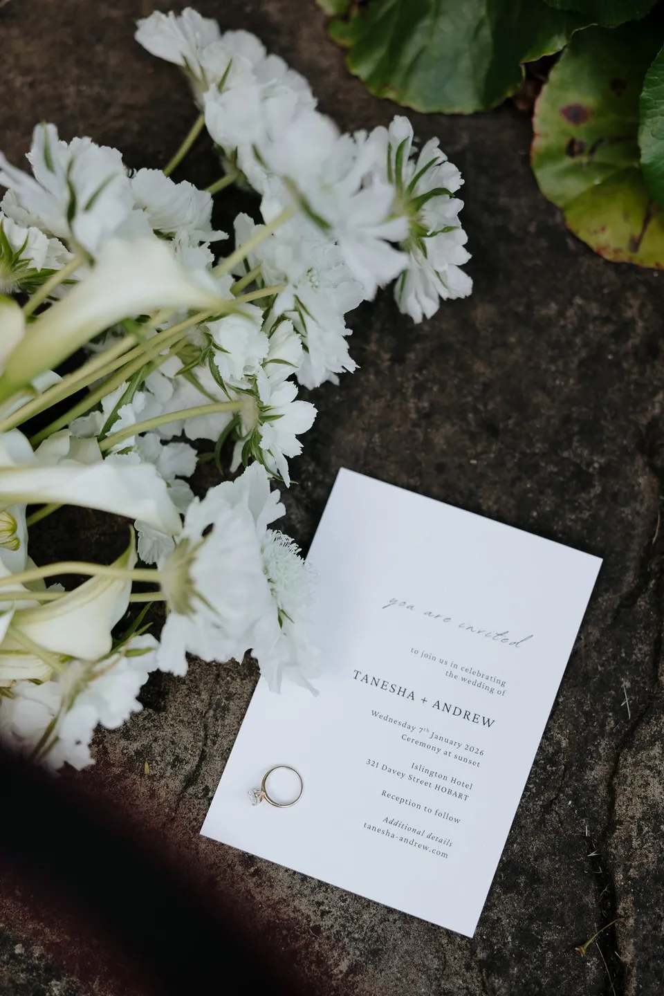 White flowers by The Romantics beside Tanesha and Andrew’s wedding invitation and rings on a stone surface, at Islington Hotel in South Hobart, Tasmania, Australia, for an Elopement Collective summer elopement. Photograph by George Bowden.