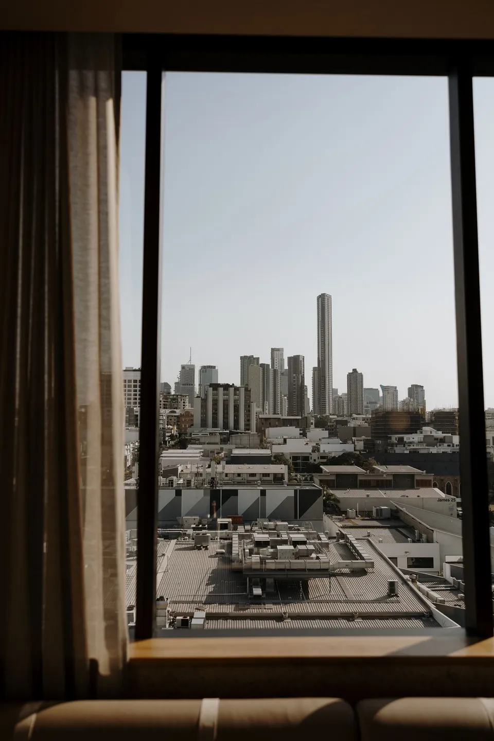 Brisbane’s skyline framed by the hotel suite window and curtains, at The Calile Hotel in Fortitude Valley, Brisbane, for an Elopement Collective elopement. Photo by George Bowden.