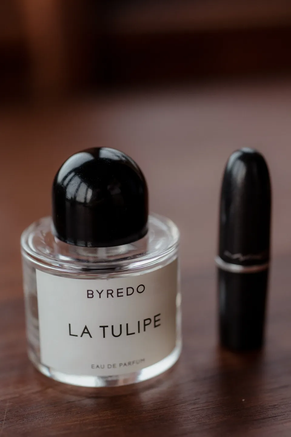 A bottle of Byredo La Tulipe perfume beside lipstick on a timber surface, at Thalia Haven on the East Coast of Tasmania, Australia, for an Elopement Collective elopement. Photograph by Joey Willis.