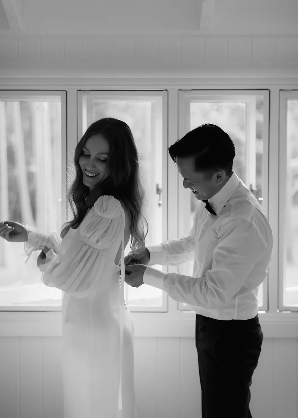 Black-and-white photograph of Elyse and Jonathan getting ready together beside the window. At Fig Tree Villa in Ewingsdale, Byron Bay, for an Elopement Collective elopement. Photograph by Bulb Creative.