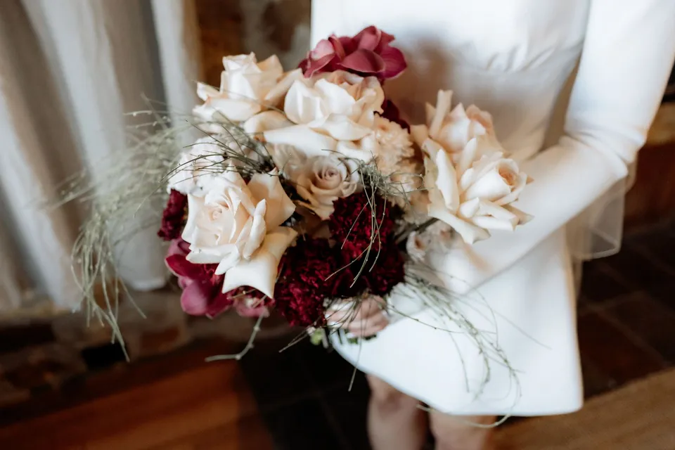 Close-up of Kaitlyn holding her cream and burgundy bouquet by The Romantics against her white dress. At Piermont Retreat on Tasmania’s east coast, for an Elopement Collective elopement. Photograph by Omar of By The Wilde.