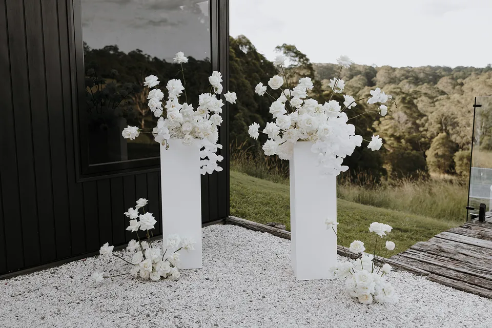 White flowers by Mrs Gibbons arranged on tall plinths and pale gravel beside the dark cabin, at Roundhill Retreat in Obi Obi, Sunshine Coast Hinterland, Queensland, for an Elopement Collective elopement. Photo by George Bowden.