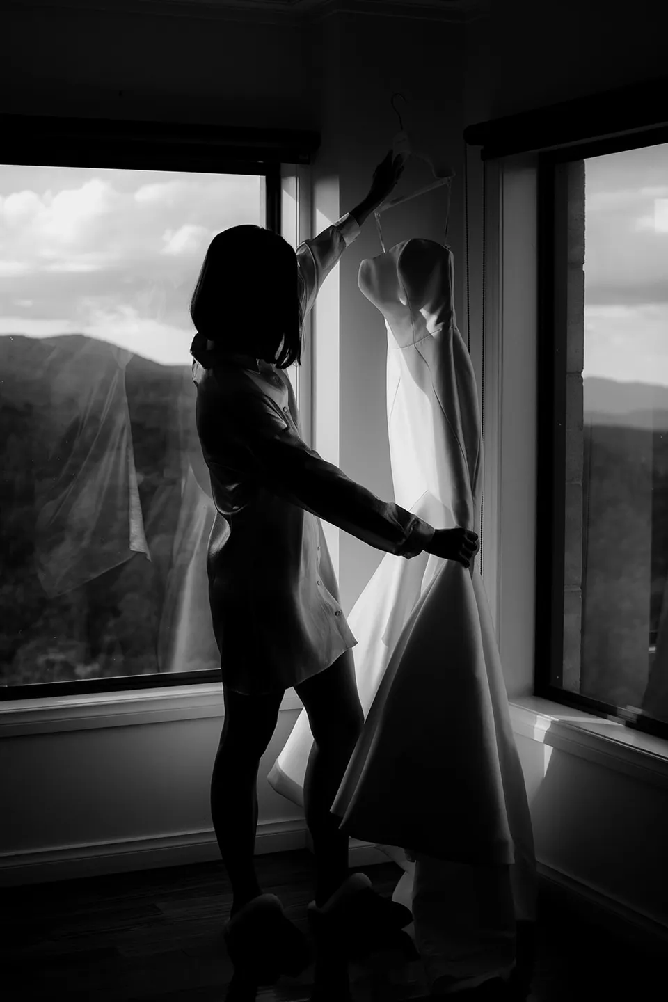 Black and white silhouette of Meg reaching for her wedding dress beside the windows. At The Keep on Tasmania’s east coast, for an Elopement Collective elopement. Photograph by Jenna Agius.