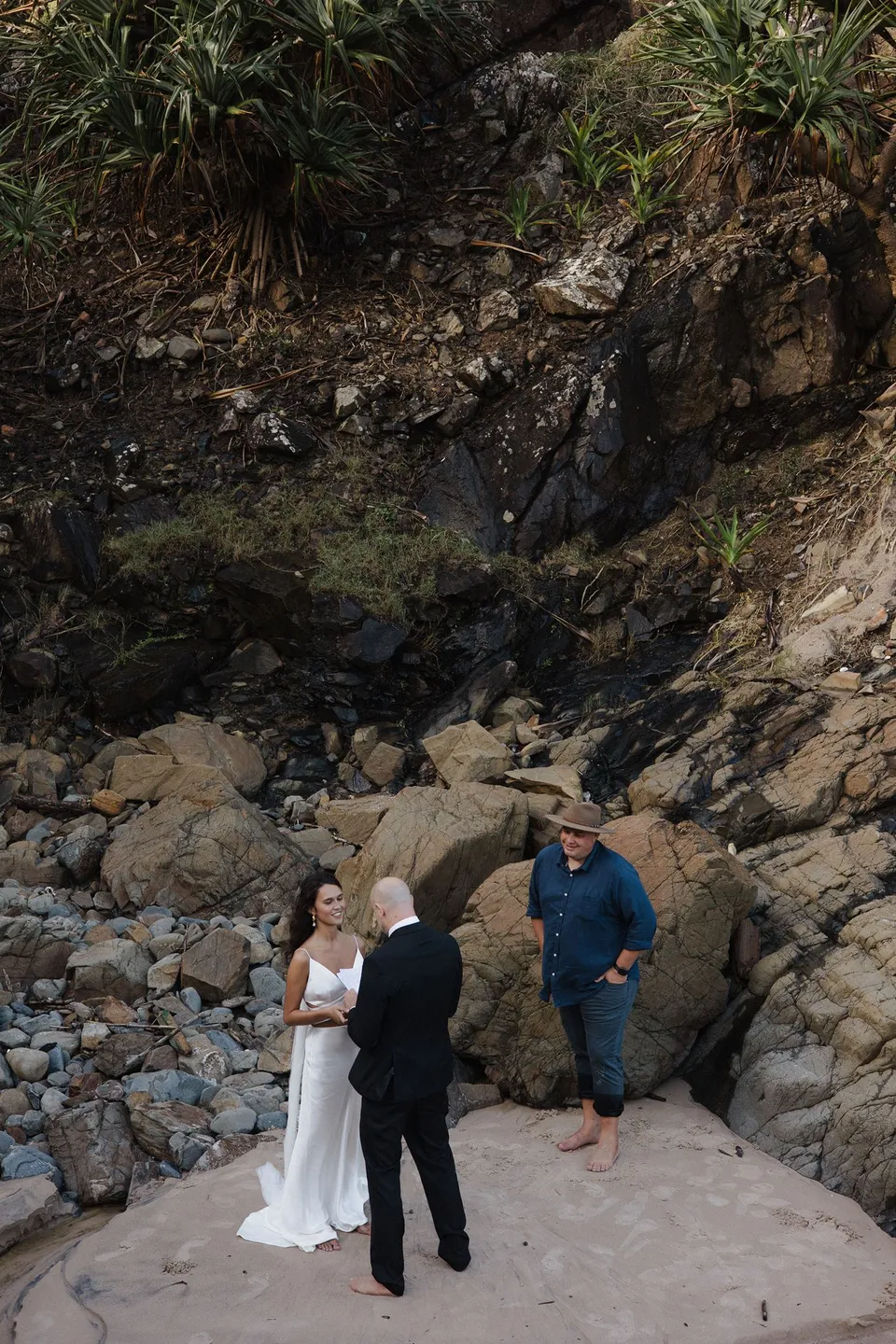 Wide view of Rachel and Dane's small ceremony beneath the rock face at Whites Beach. At Whites Beach, Broken Head, near Byron Bay, for an Elopement Collective elopement. Photograph by Jason Lucas.