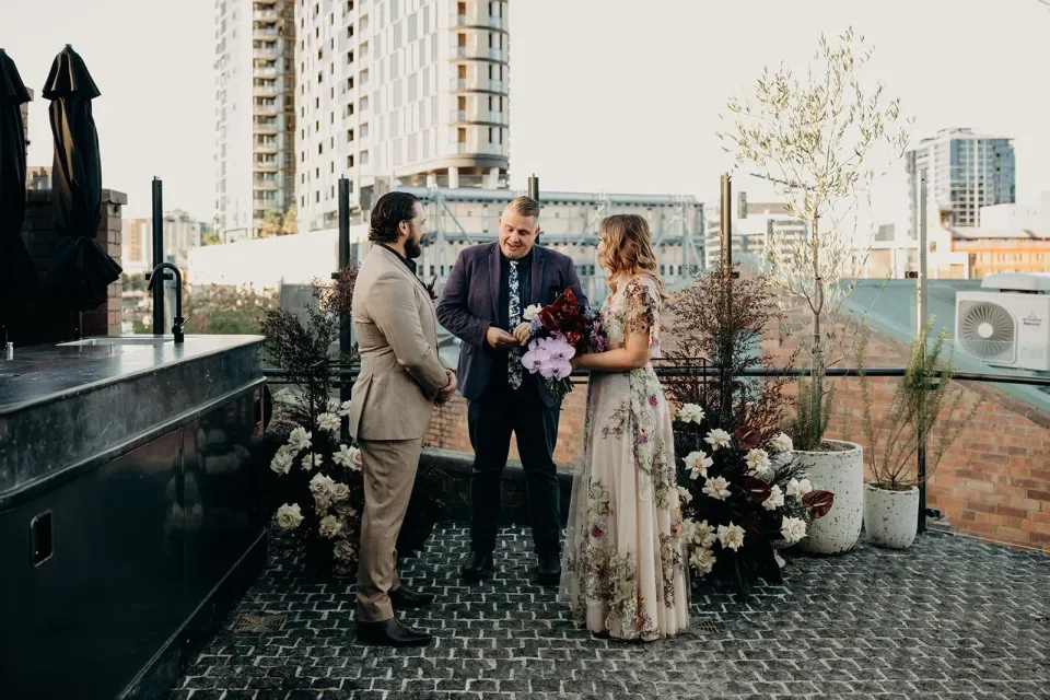 Celebrant Josh Withers leading Victoria and Daniel’s rooftop ceremony between arrangements by Mrs Gibbons Flowers, at Agnes Restaurant in Fortitude Valley, Brisbane, for an Elopement Collective elopement. Photo by Tessa Shannon.