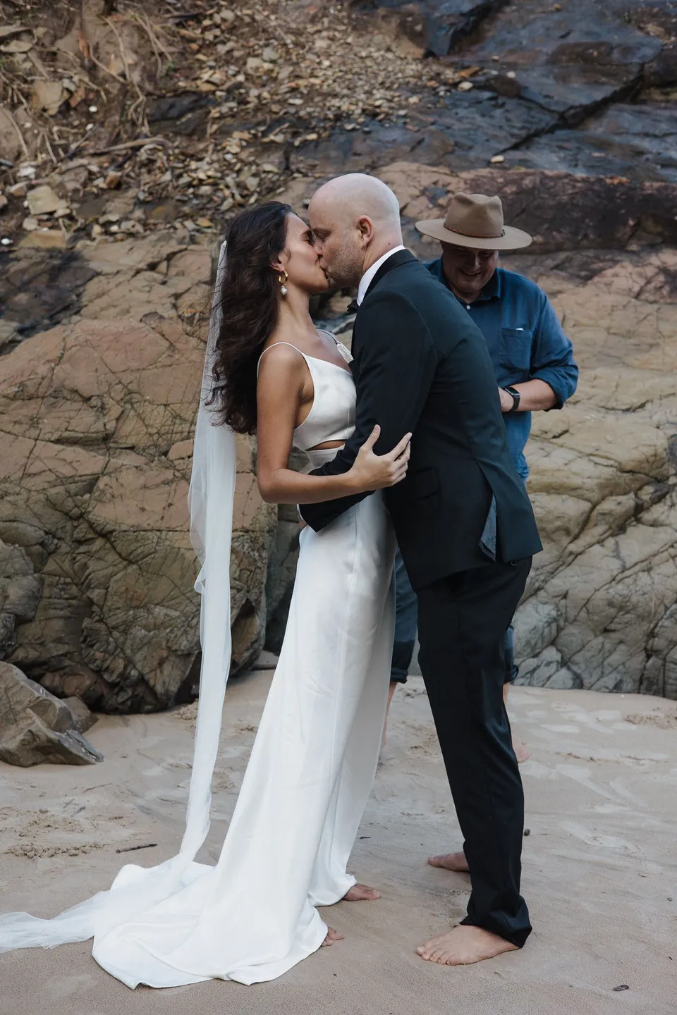 Rachel and Dane kissing beside the rocks with celebrant Josh Withers behind them. At Whites Beach, Broken Head, near Byron Bay, for an Elopement Collective elopement. Photograph by Jason Lucas.