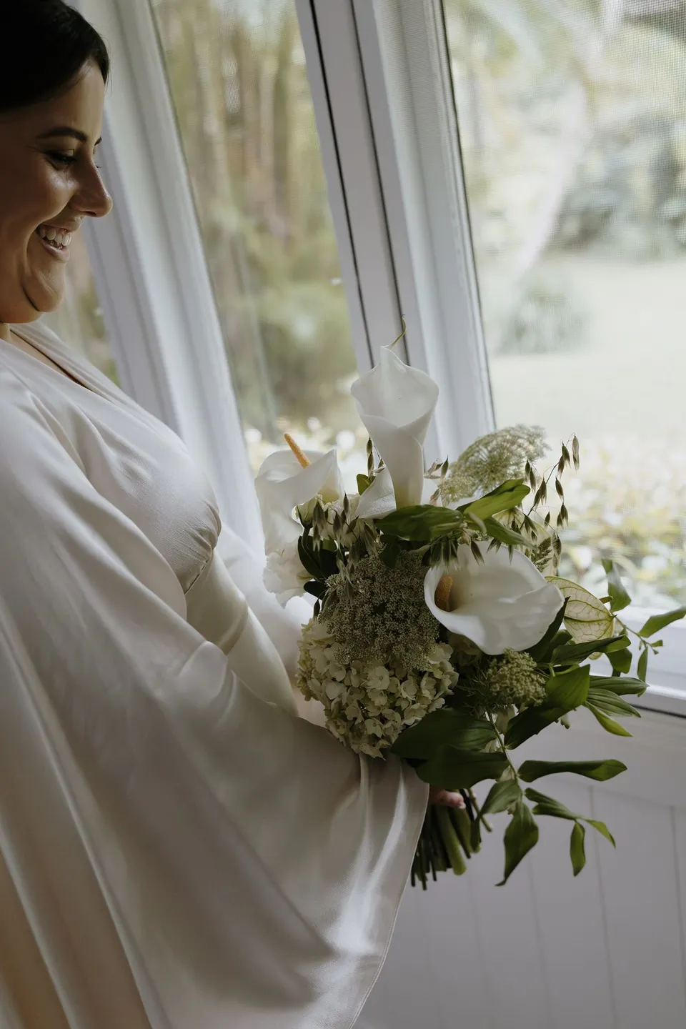 Laura holding her white and green bouquet by The French Petal beside a window. At Fig Tree Villa in Ewingsdale, Byron Bay, for an Elopement Collective elopement. Photograph by George Bowden.
