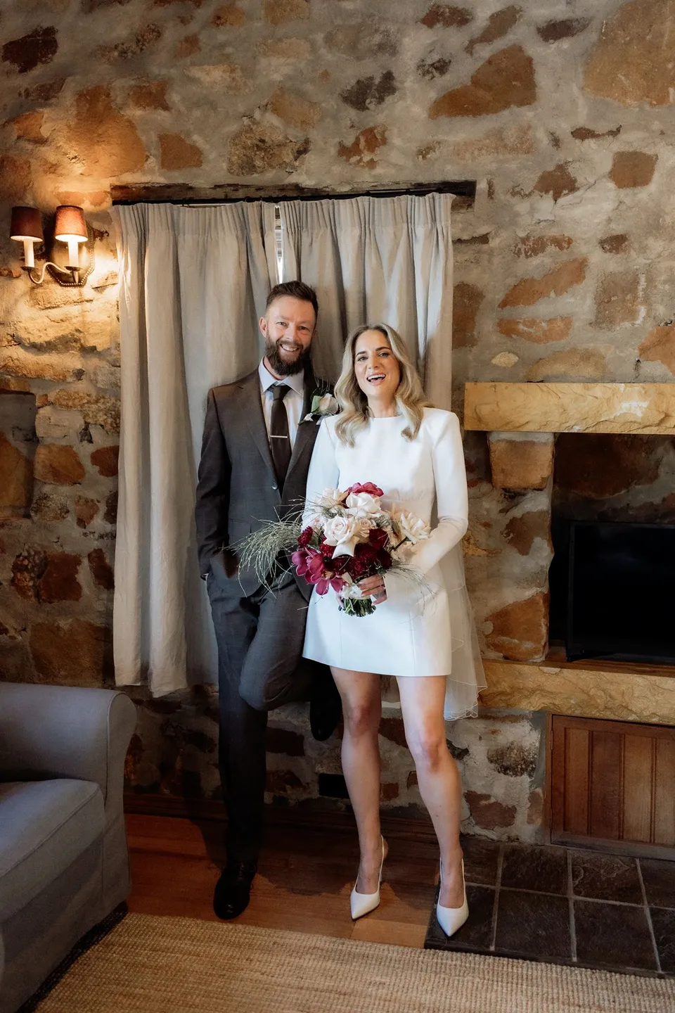 Kaitlyn and Russell smiling together inside the stone cottage, with her bouquet by The Romantics. At Piermont Retreat on Tasmania’s east coast, for an Elopement Collective elopement. Photograph by Omar of By The Wilde.
