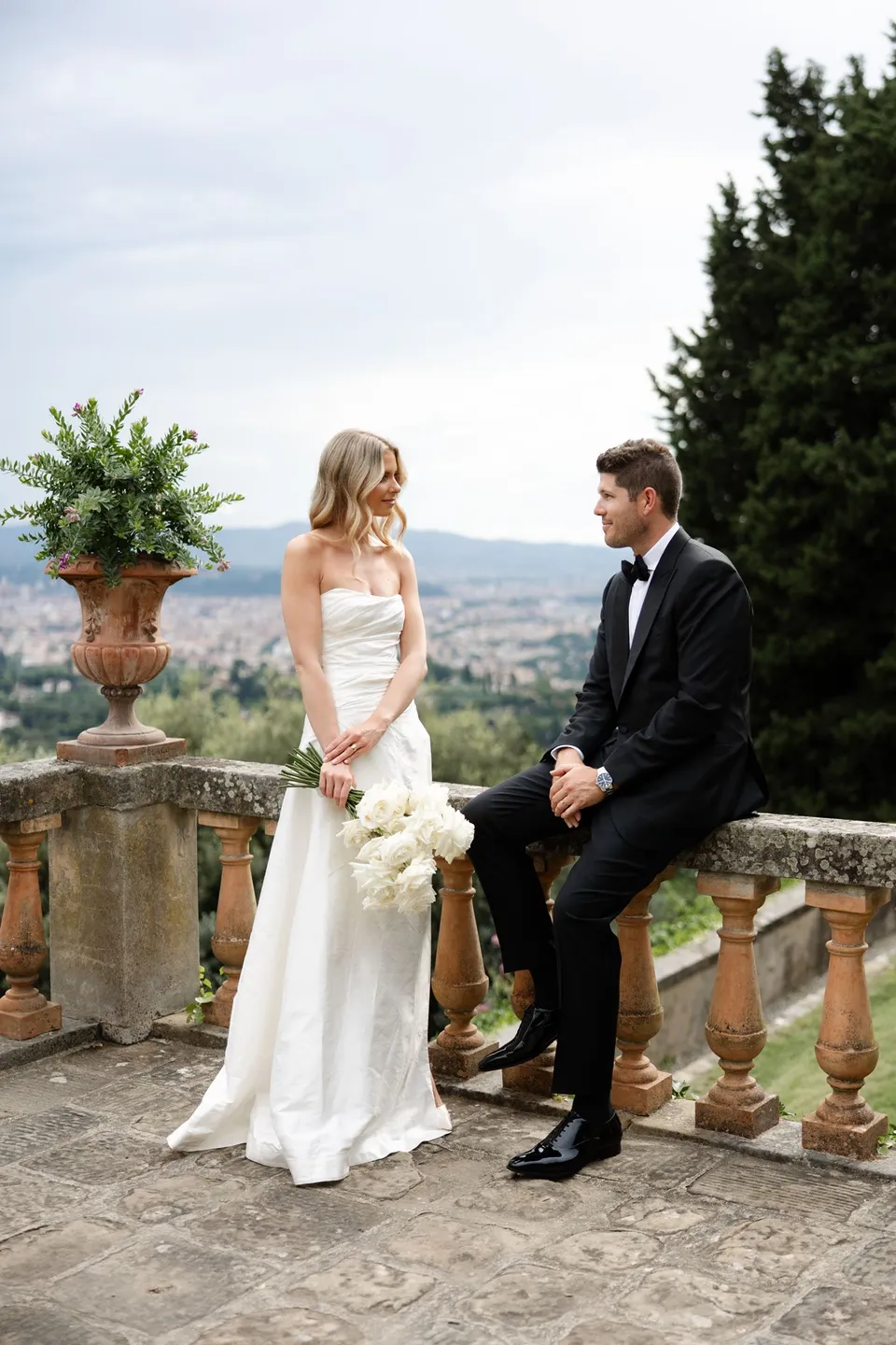Matthew sitting on the balustrade as Holly stands beside him with her white bouquet by Le Francesche Flower Designers, at Villa San Michele in Fiesole above Florence, Tuscany, Italy, for an Elopement Collective elopement. Digital photograph by Khara from House of Lucie.