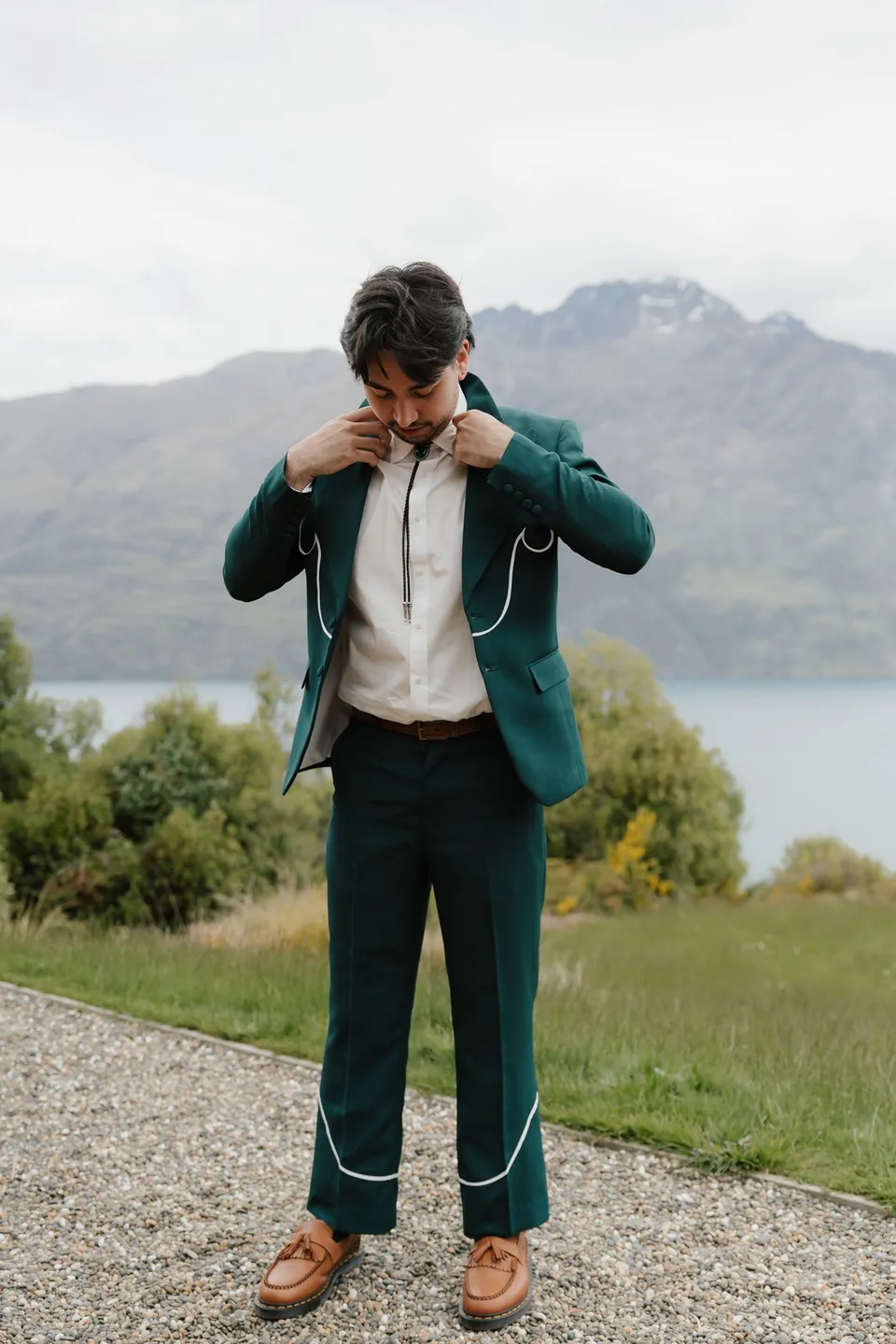 Adrian adjusting his bolo tie in a deep green suit with white piping, with the lake and mountains behind him, at their alpine Airbnb in Ben Lomond, Queenstown, New Zealand, for an Elopement Collective Queenstown elopement. Photograph by Bulb Creative.