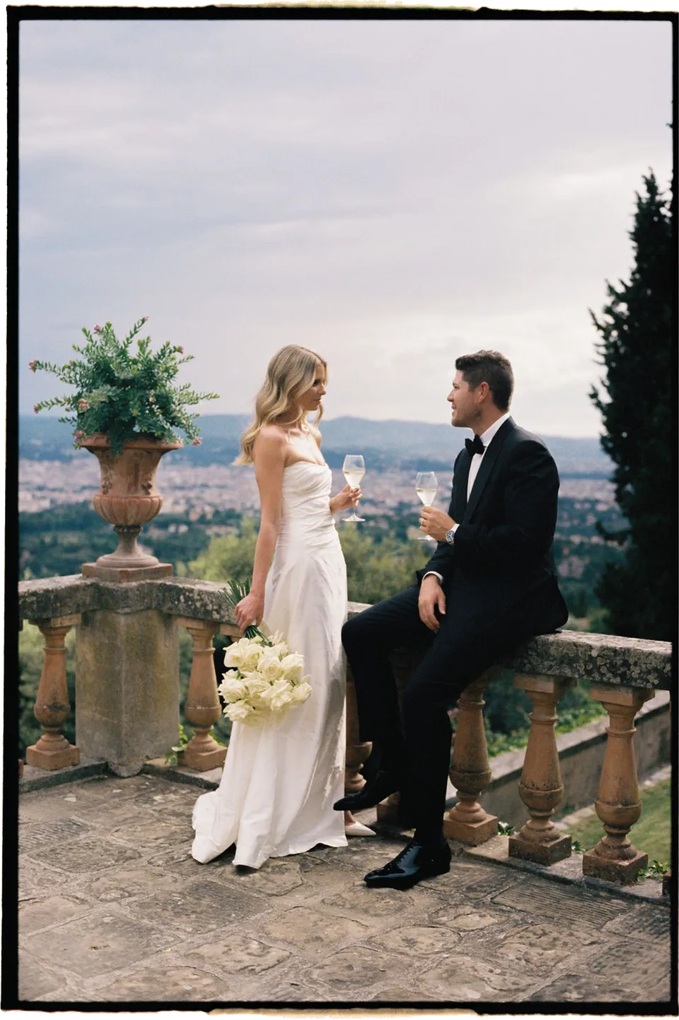 Holly holding her white bouquet by Le Francesche Flower Designers beside Matthew on the stone balustrade, at Villa San Michele in Fiesole above Florence, Tuscany, Italy, for an Elopement Collective elopement. Film photograph by Khara from House of Lucie.