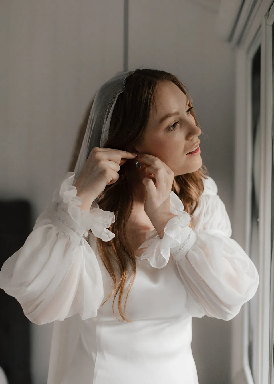 Elyse fastening an earring beside the window. At Fig Tree Villa in Ewingsdale, Byron Bay, for an Elopement Collective elopement. Photograph by Bulb Creative.