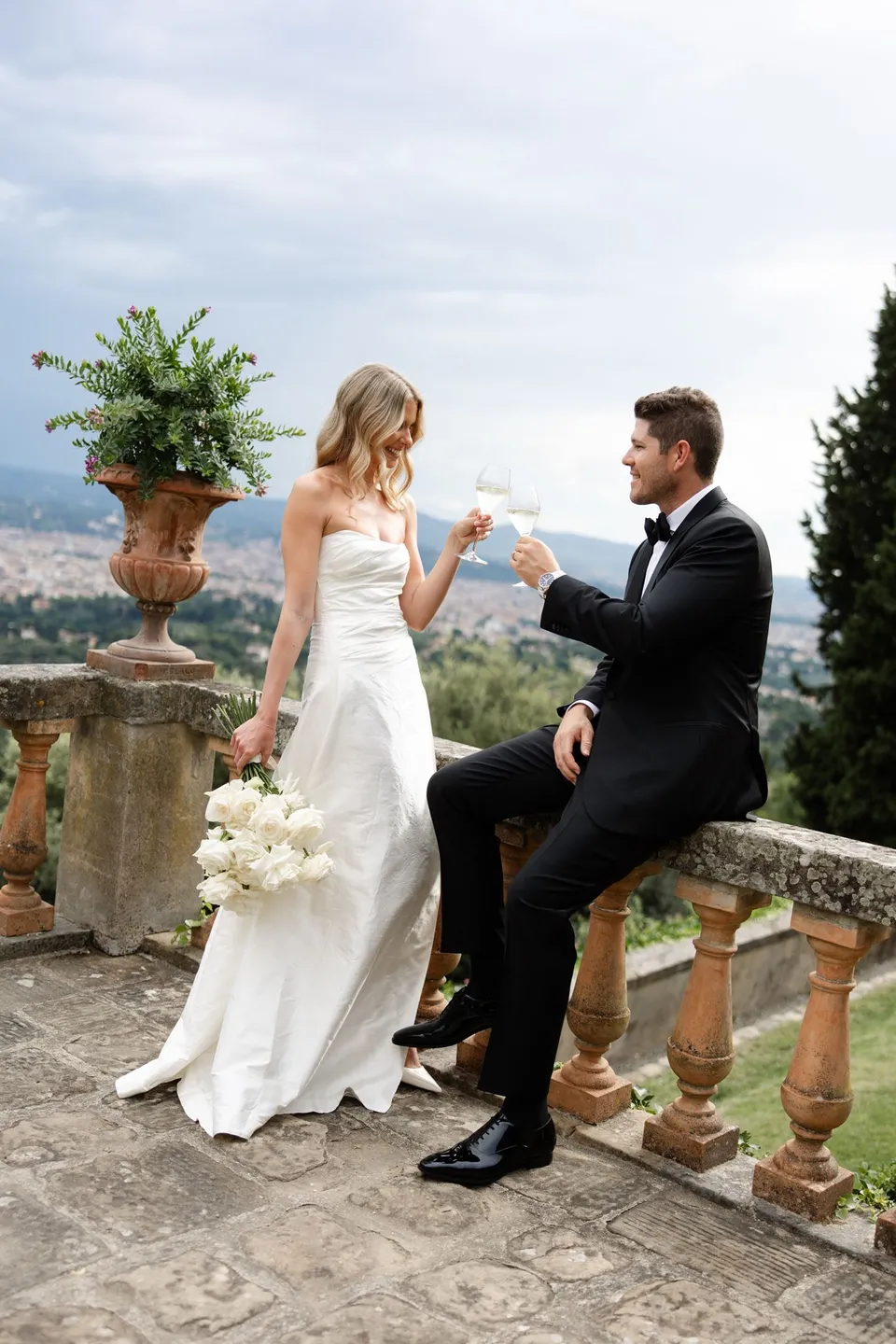 Holly and Matthew smiling at each other with glasses of wine on the terrace, beside her white bouquet by Le Francesche Flower Designers, at Villa San Michele in Fiesole above Florence, Tuscany, Italy, for an Elopement Collective elopement. Digital photograph by Khara from House of Lucie.